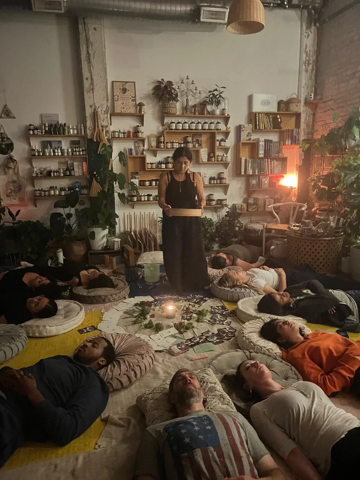 A woman standing in a dimly lit room with a group of people lying on the floor in a circle, participating in a guided meditation or relaxation session. The room is decorated with plants, shelves filled with jars and books, and ambient lighting, creating a cozy and tranquil atmosphere.