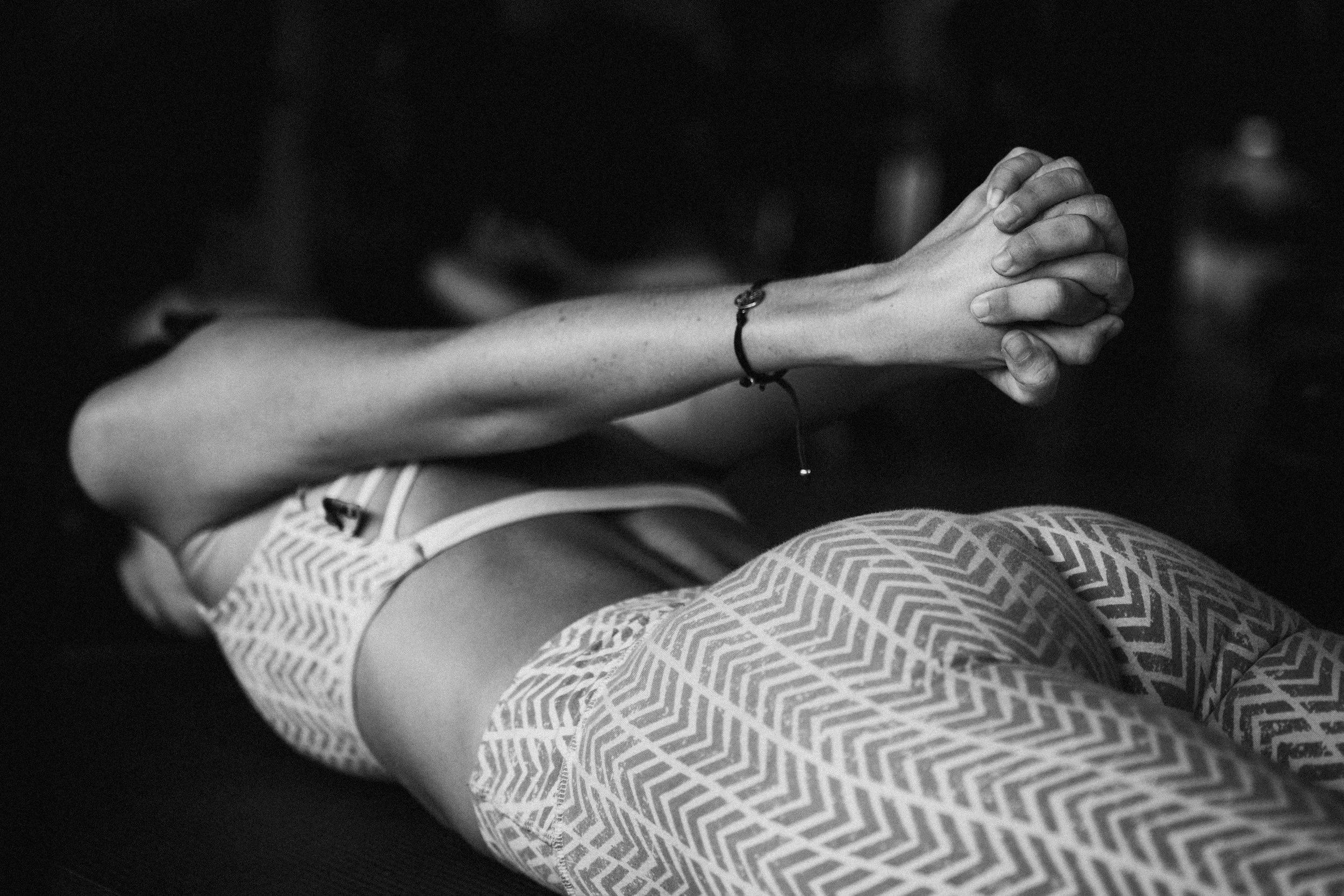 A person clasping their hands in a prayer or meditative pose, lying on their side on a yoga mat, wearing patterned leggings and a tank top.