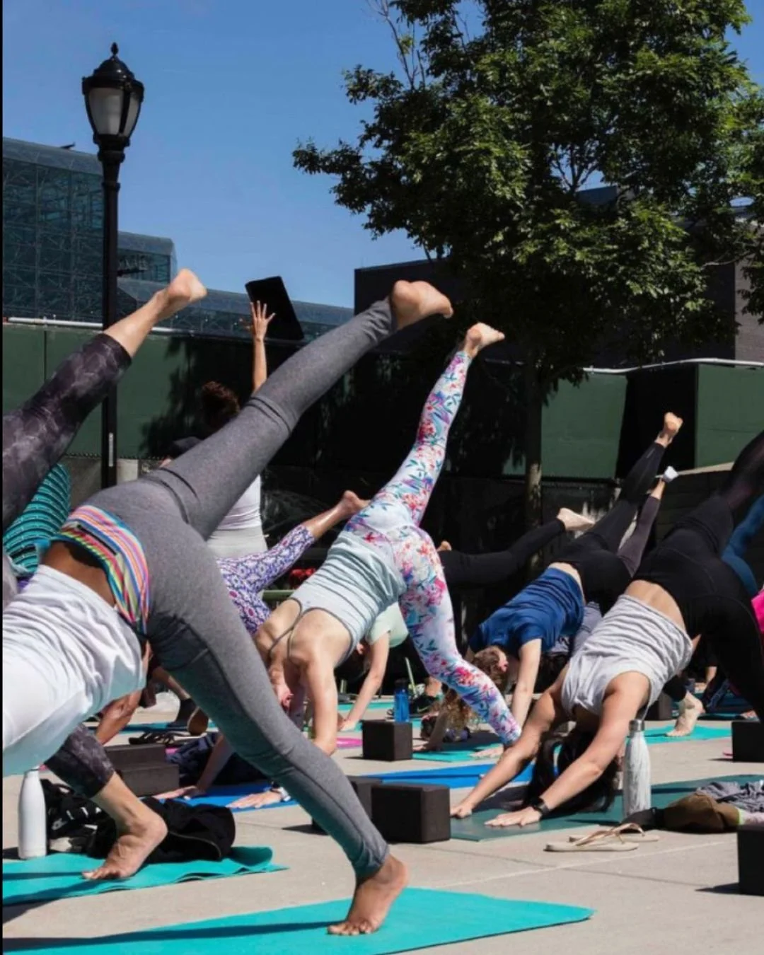 People practicing yoga outdoors on mats, performing downward dog poses with one leg raised, during a sunny day in a public space with trees, a lamp post, and modern buildings in the background.