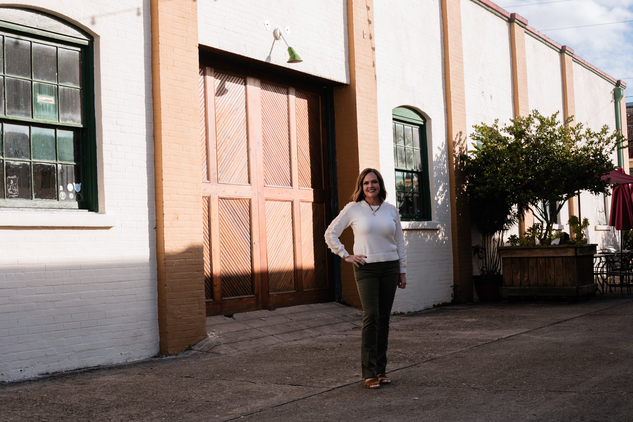 A woman standing outside in front of a brick building with wooden doors and windows, smiling and posing with one hand on her hip.