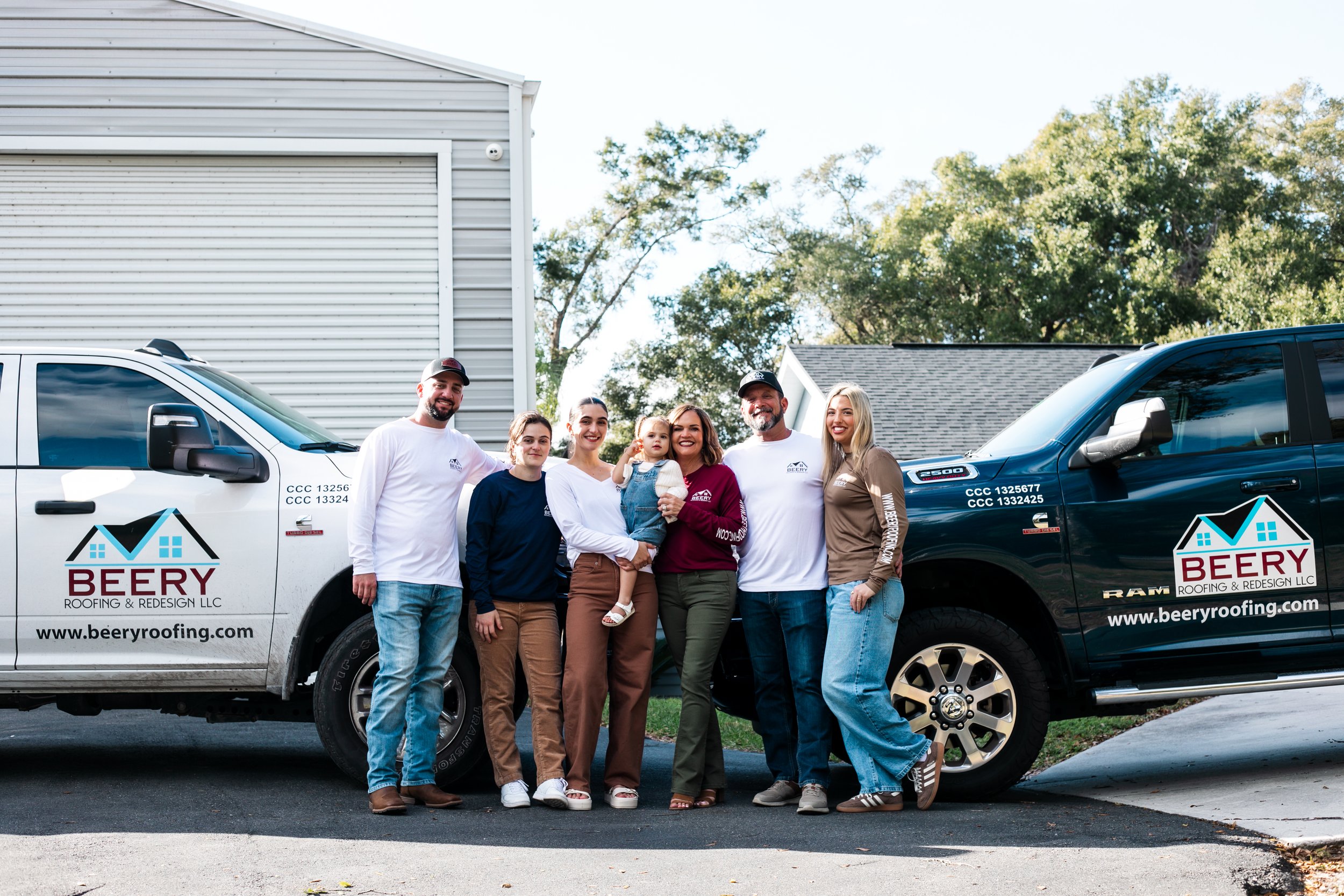 Group of seven people standing outdoors between two company trucks, smiling, with trees and a building in the background.