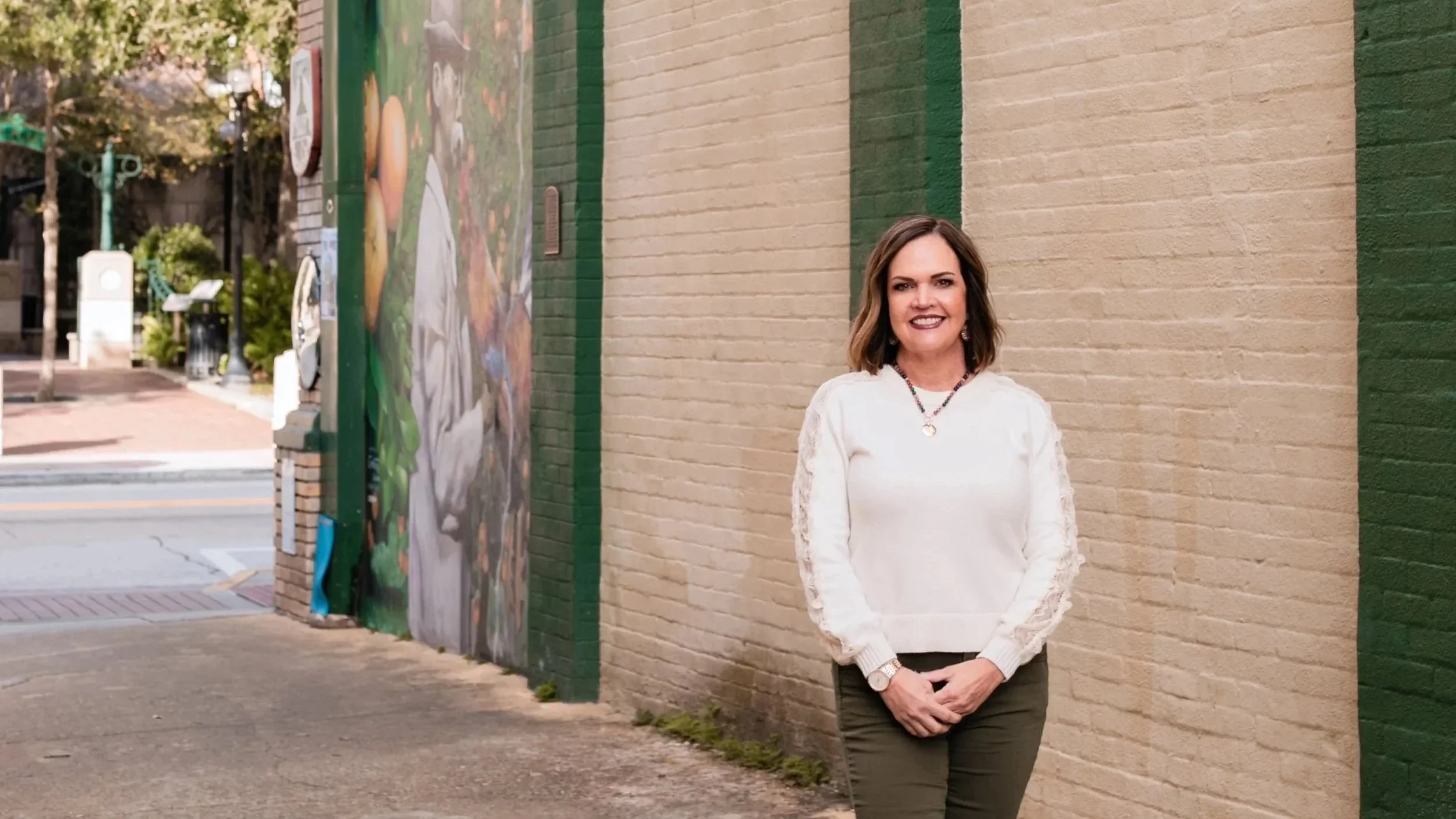 A woman with shoulder-length brown hair, wearing a white sweater with lace sleeves, dark green pants, a watch, and a necklace, standing against a beige brick wall with green vertical lines on a city sidewalk.