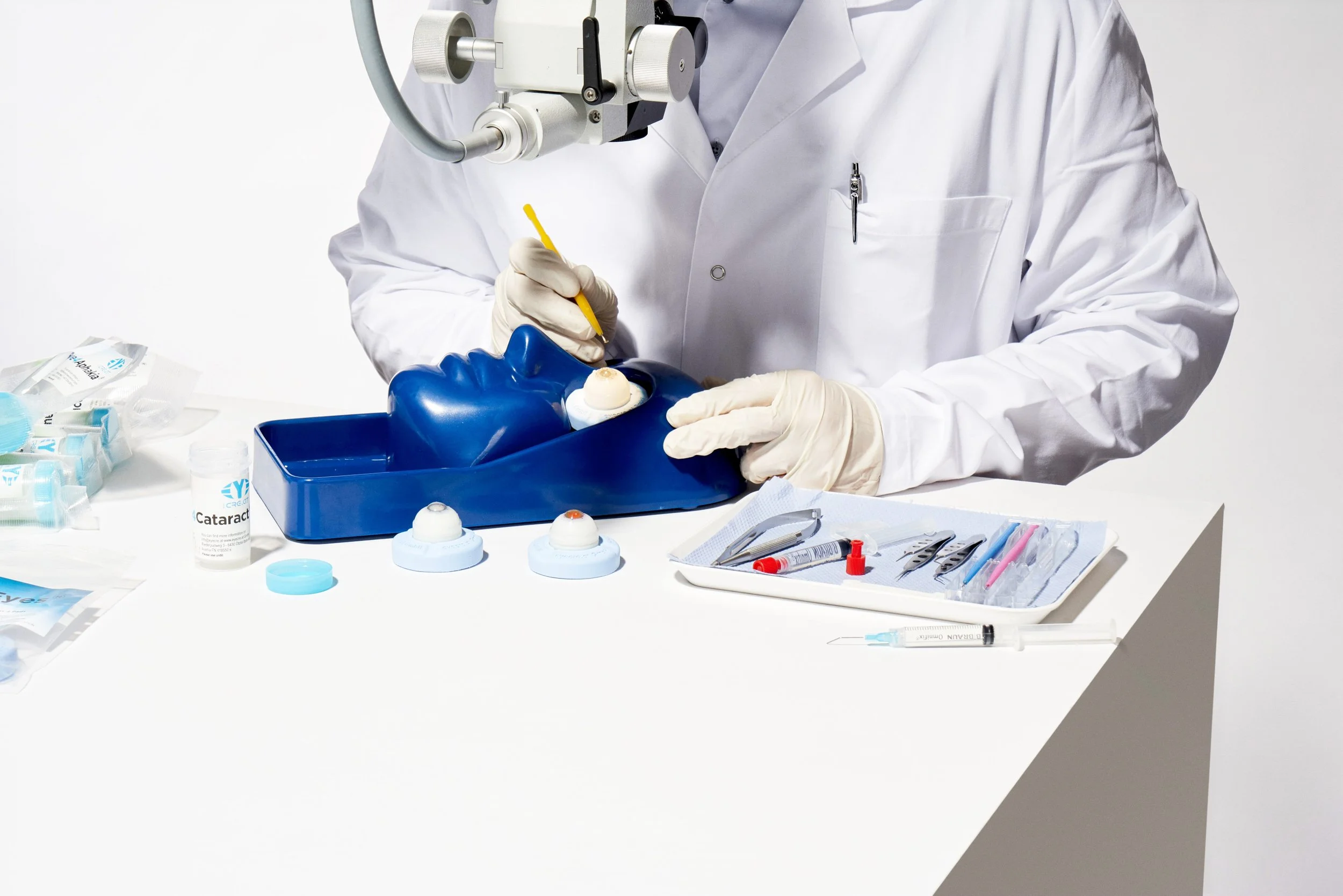 A medical professional in a white lab coat and gloves performing a simulated eye surgery on a practice model, surrounded by surgical tools and supplies.