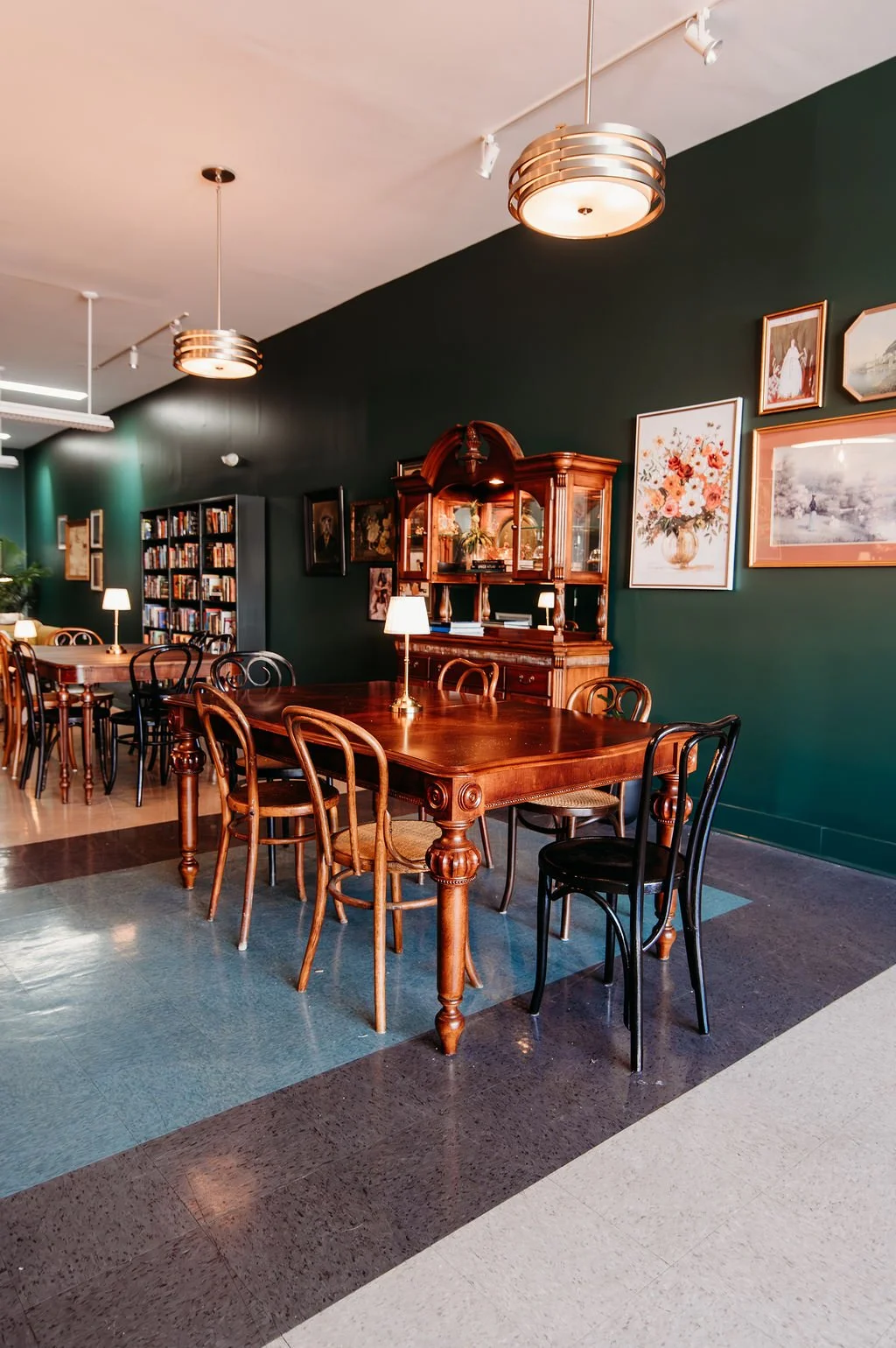 A cozy, vintage-style room with wooden tables and chairs, green walls decorated with framed artwork, and a bookshelf, illuminated by modern pendant lighting and small table lamps.