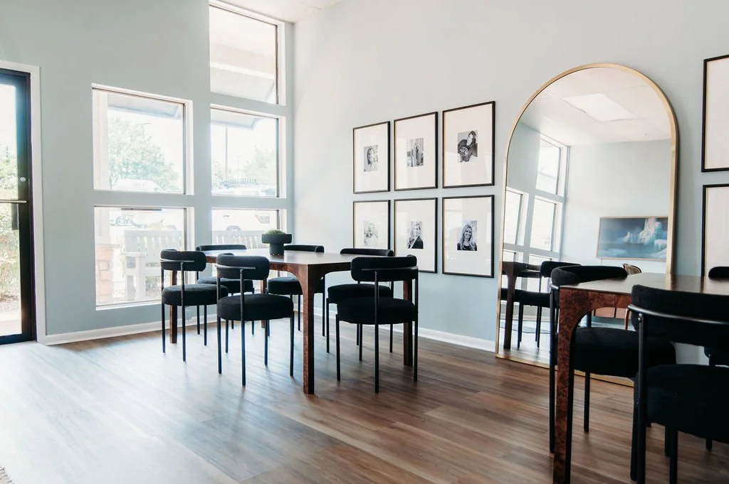 A bright co-working office area with large windows, a wooden table, black chairs, framed artwork on the wall, and a large mirror reflecting the room.