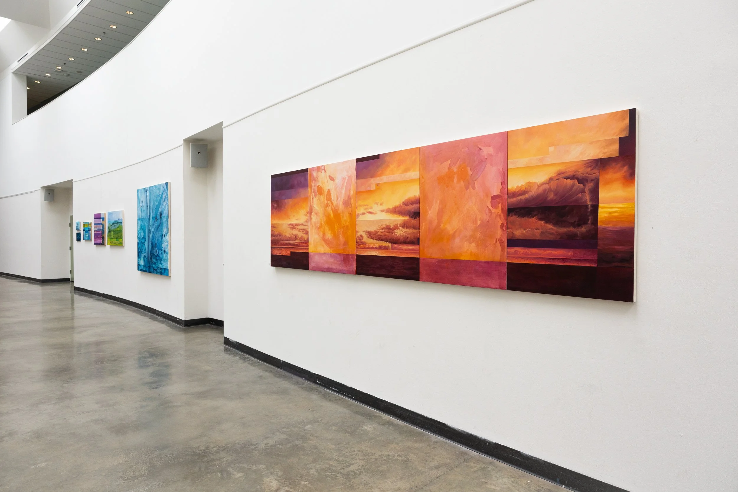 Art gallery with multiple colorful paintings hanging on a white wall, showing abstract and landscape scenes, in a spacious, well-lit area with polished concrete floors.