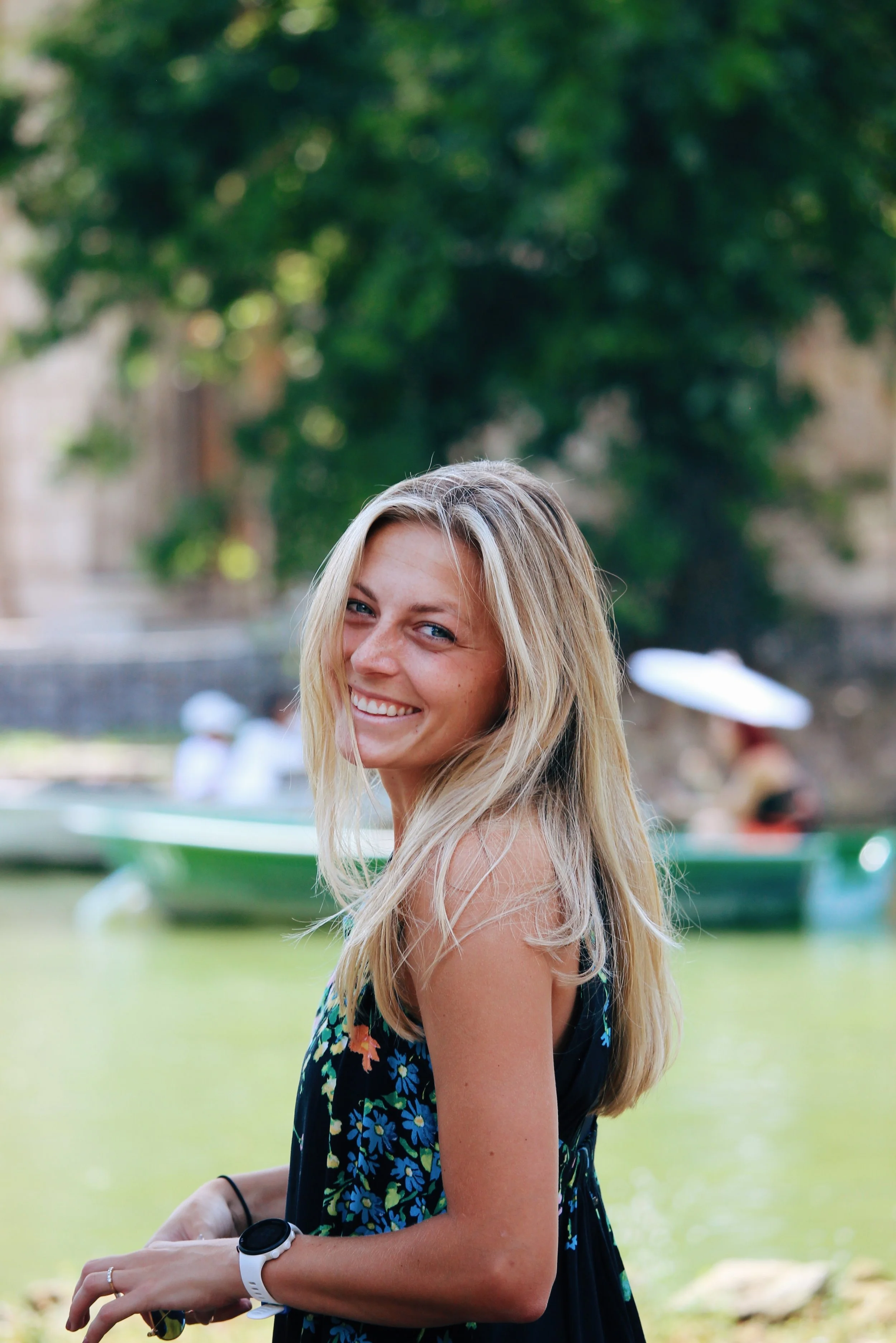 A young woman with long blonde hair and blue eyes smiling outdoors near water, with a blurred background of boats and trees.