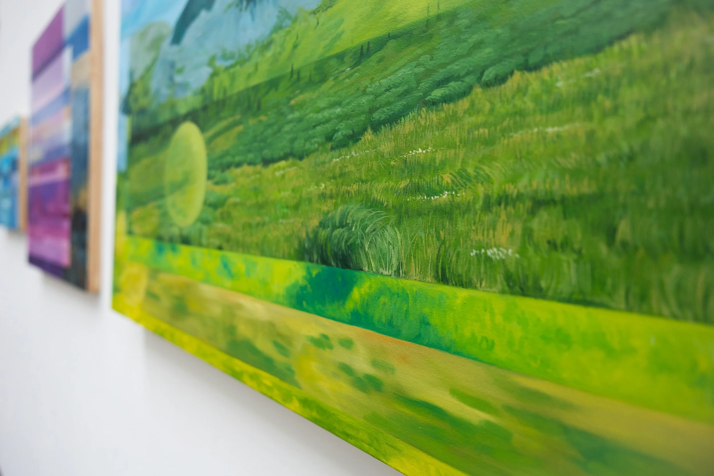 Close-up of a landscape painting on a wall, featuring a green field and blue sky in a bright, colorful scene.