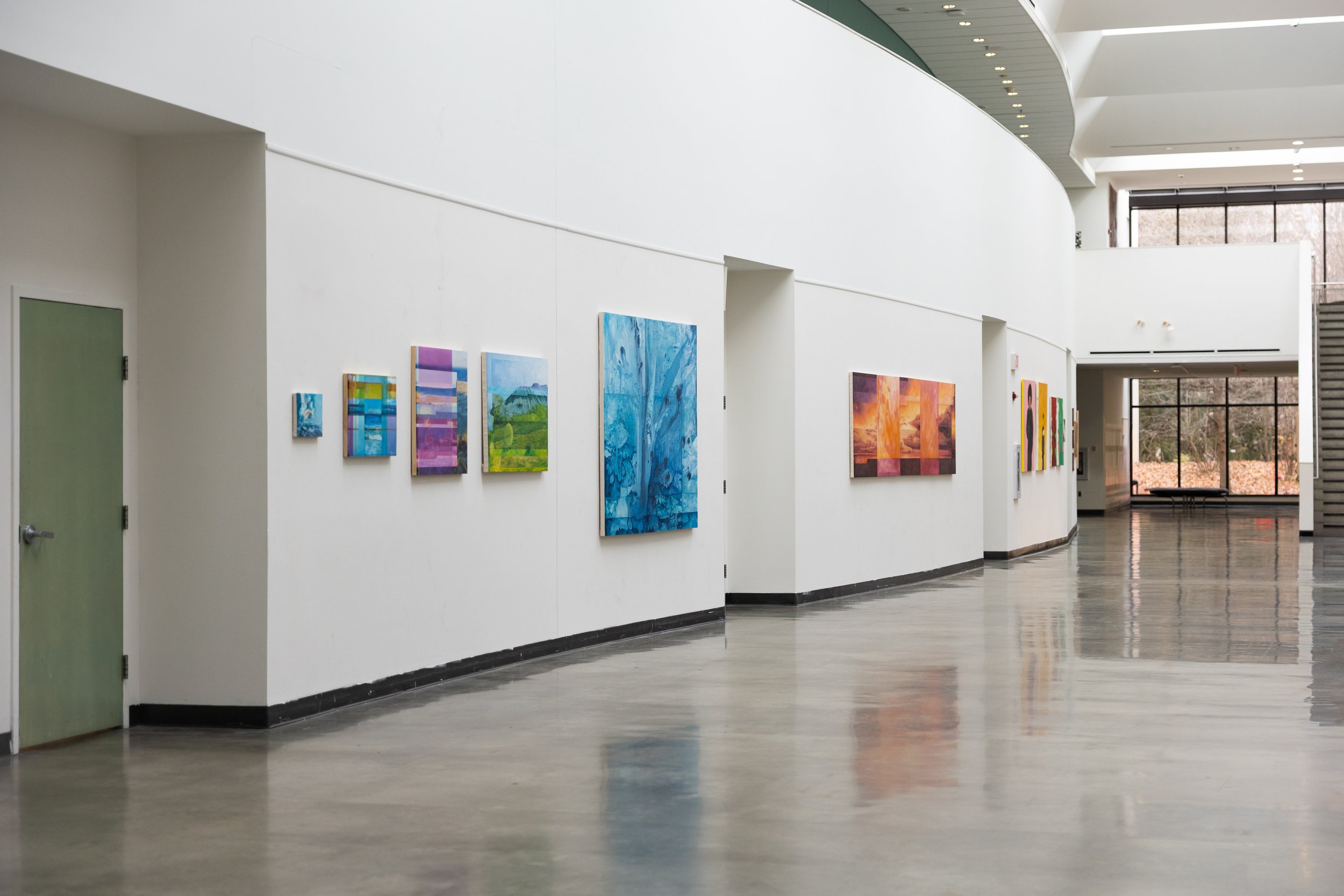 Art gallery with colorful paintings displayed on white walls, large windows letting in natural light, and a polished concrete floor.