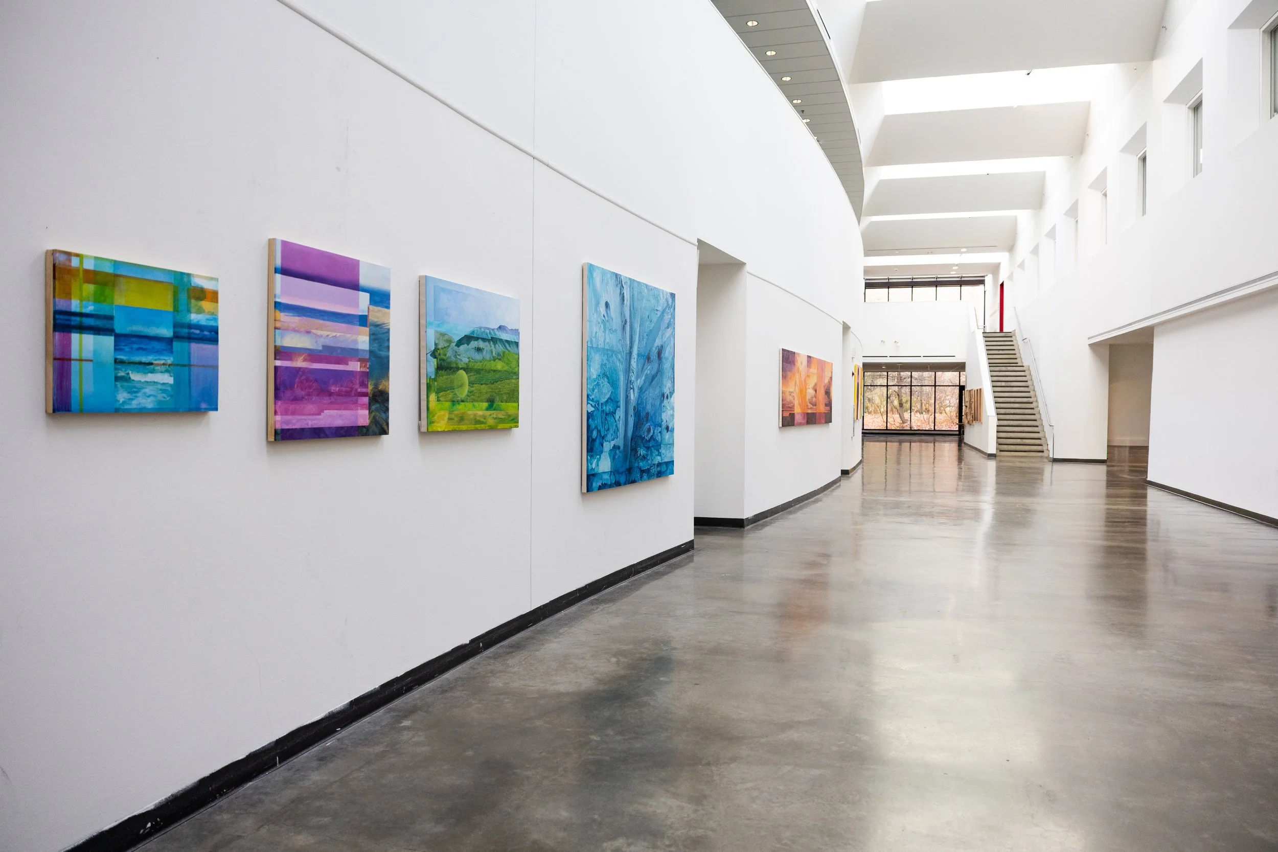 An art gallery with multiple colorful paintings on white walls and a polished concrete floor. A staircase is visible on the right, and large windows allow natural light to fill the space.