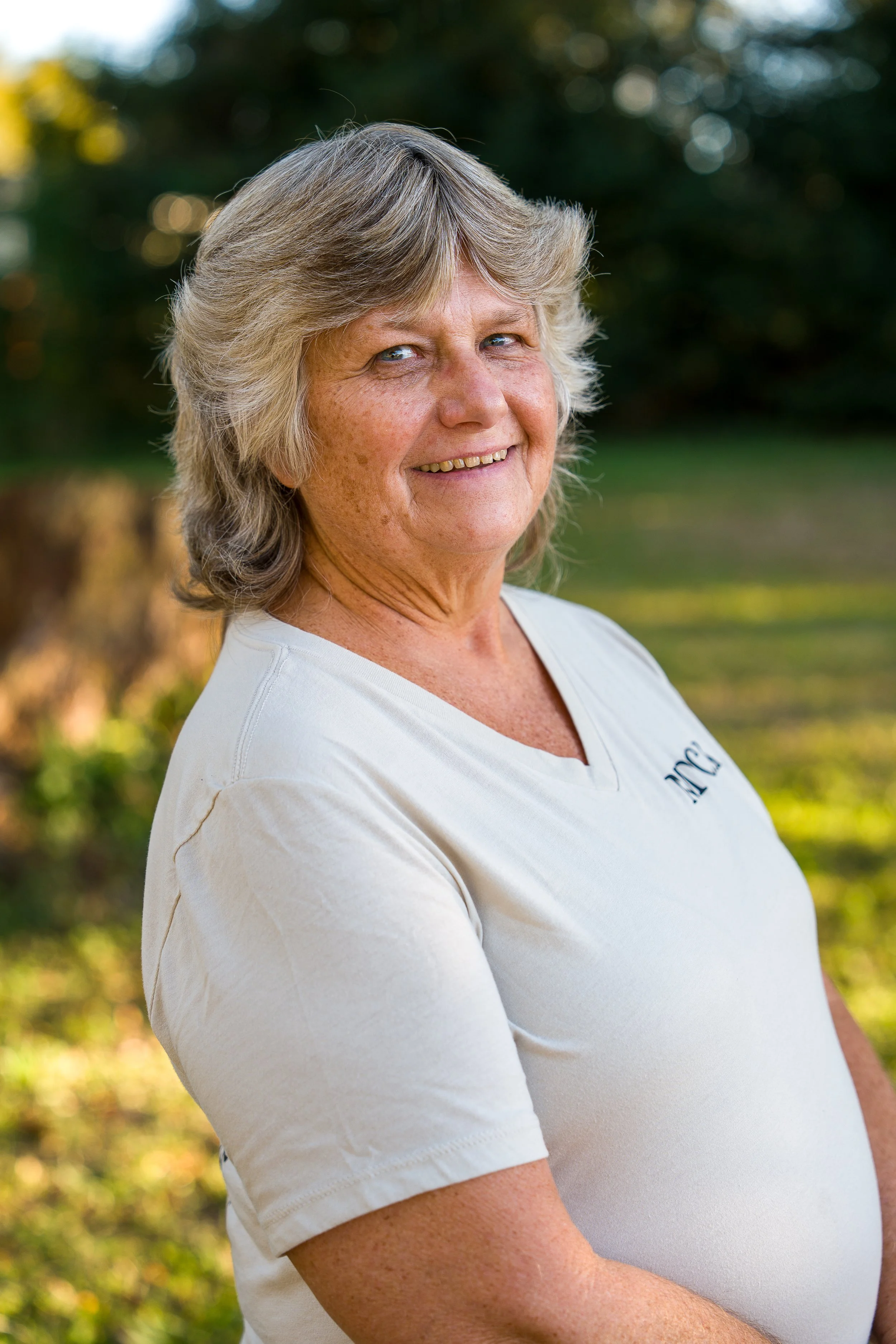 A woman with short gray hair and sunglasses taking a selfie at the beach, with a pier in the background and the ocean shining under a clear blue sky.