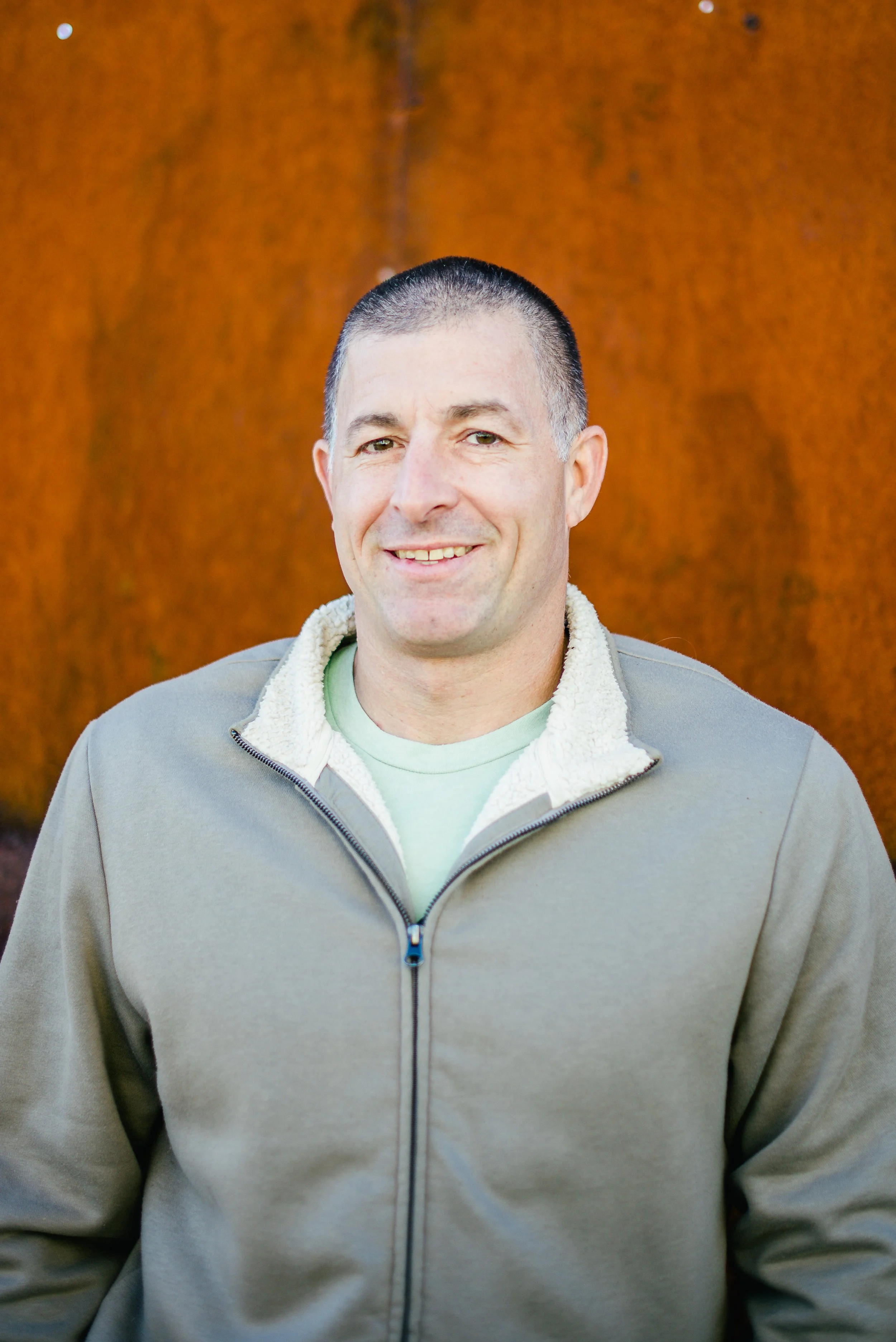 A man with short dark hair and a slight smile, wearing a beige zip-up jacket with a cream fleece collar, standing outdoors in front of a large rust-colored metal wall.