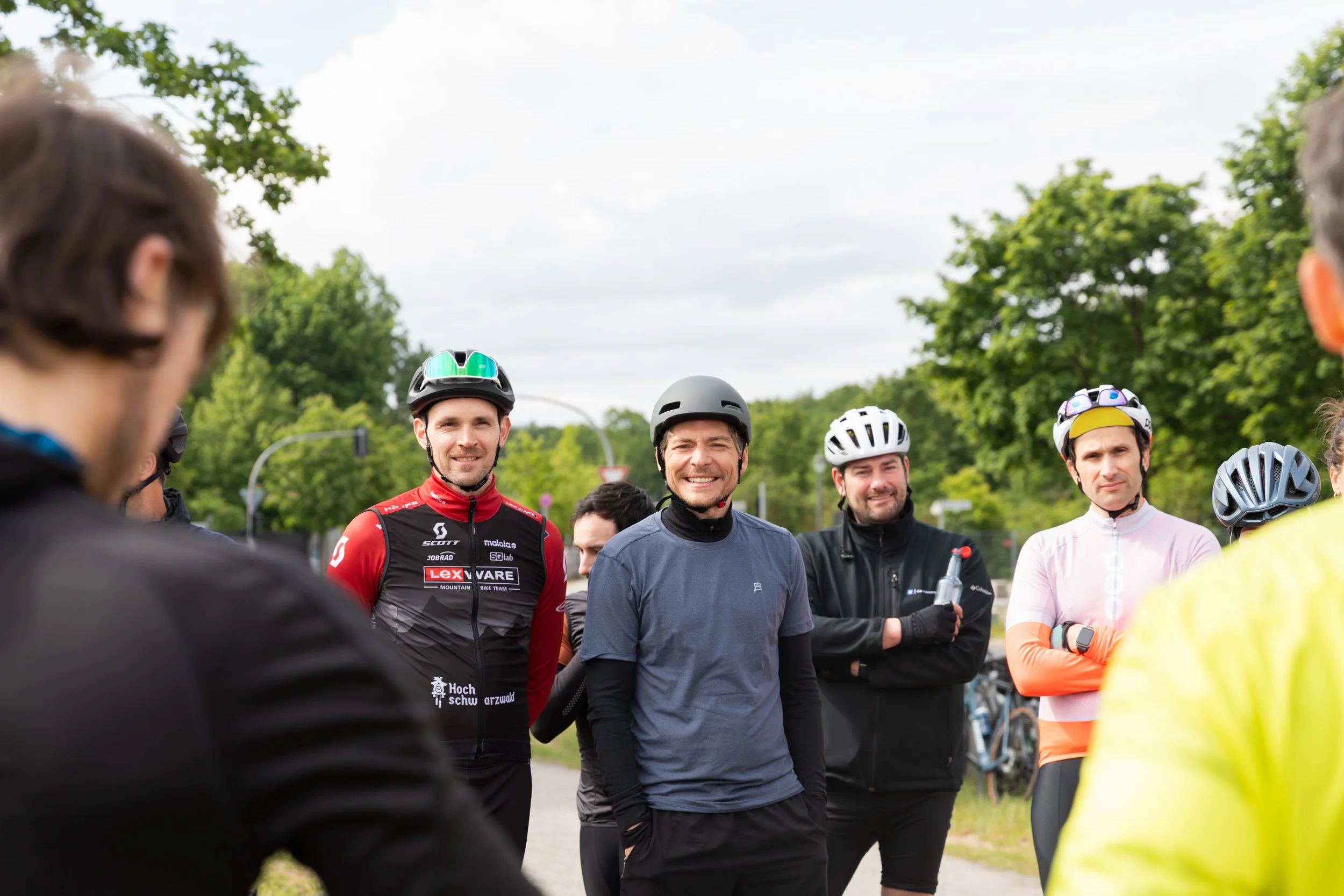 Gruppe von Radfahrern mit Helmen,die im Freien stehen, im Hintergrund Bäume und ein grauer Himmel.