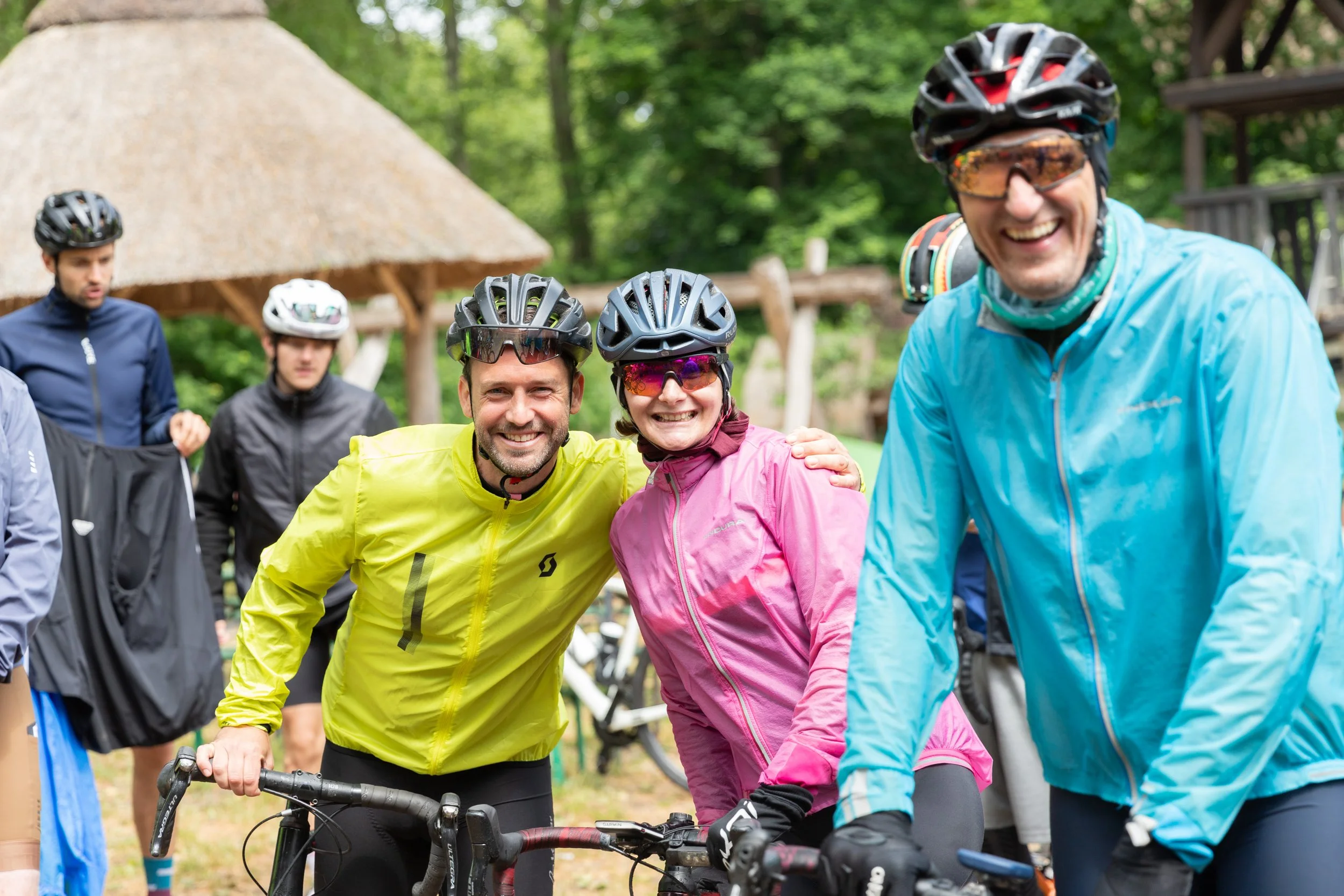 Gruppe von Fahrradfahrern in bunten Jacken und Helmen, lachend und in der Natur, vor einem Holzunterstand.