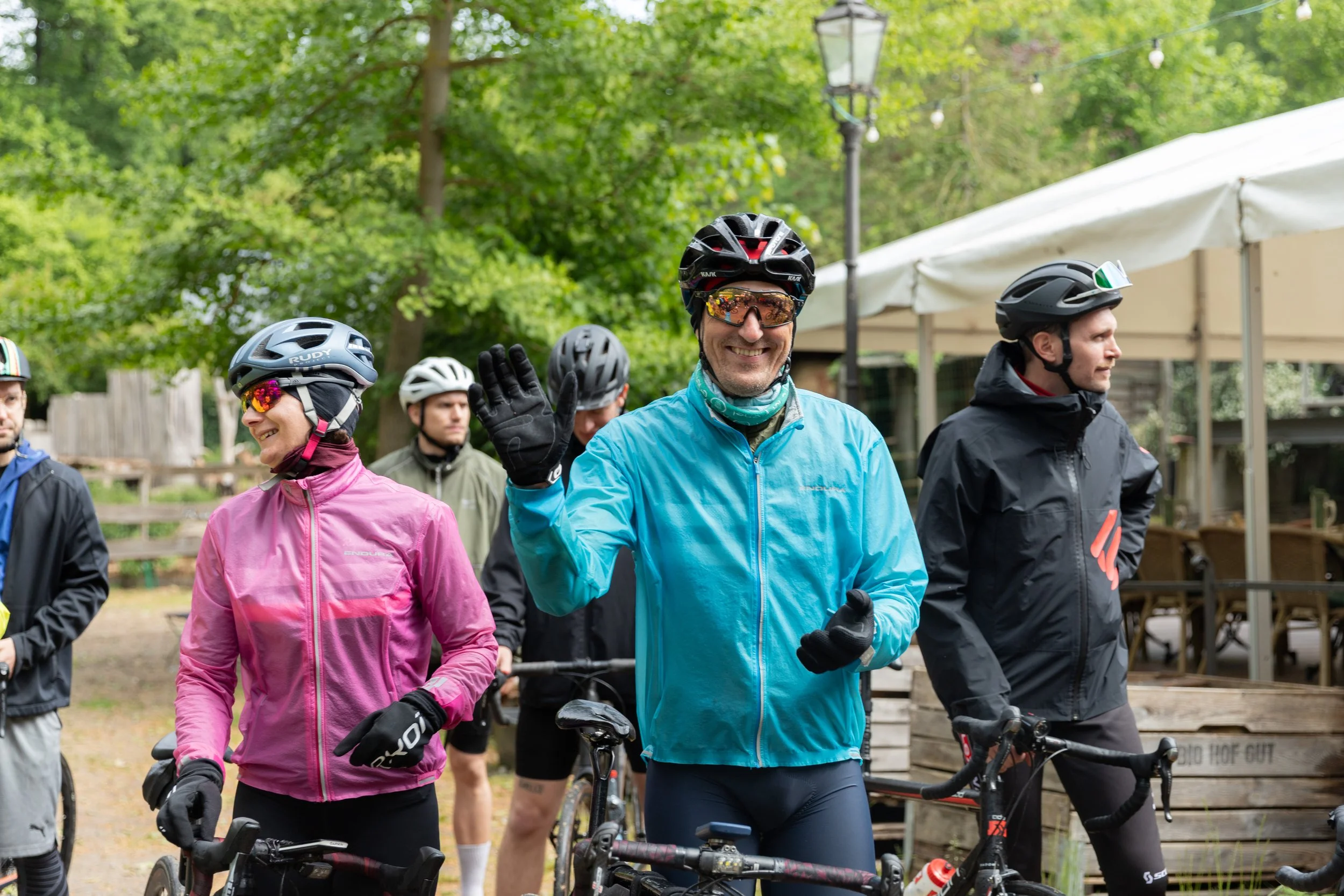 Eine Gruppe von Mountainbikern, darunter ein Mann im blauen Jacke, lächelt und winkt, während sie mit ihren Fahrrädern im Freien stehen, umgeben von Bäumen und einer Zeltsitzgruppe.