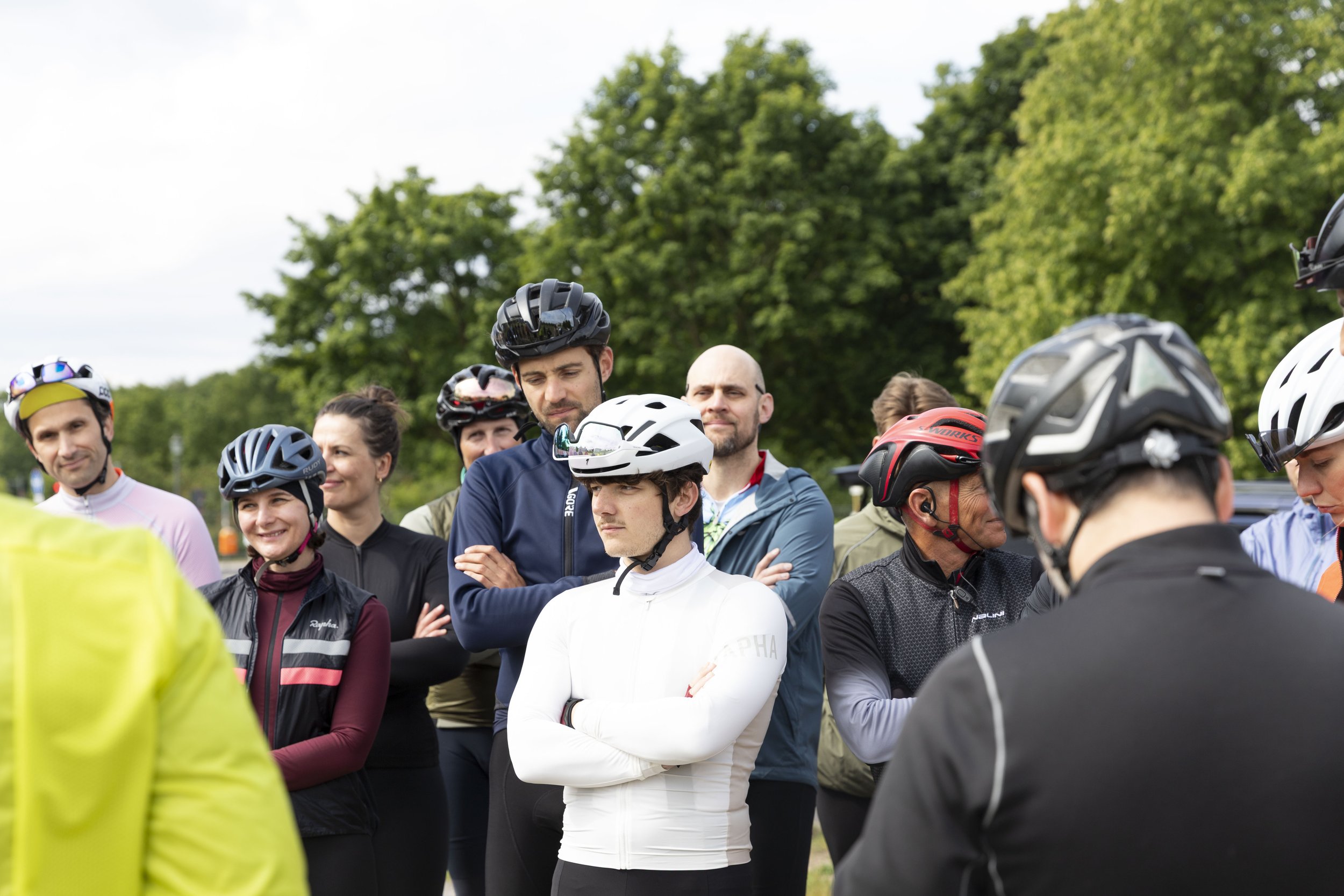 Gruppe von Fahrradfahrern im Gespräch, alle tragen Fahrradhelme und sportliche Kleidung, im Hintergrund Bäume, vermutlich bei einem Treffen oder einer Veranstaltung im Freien.