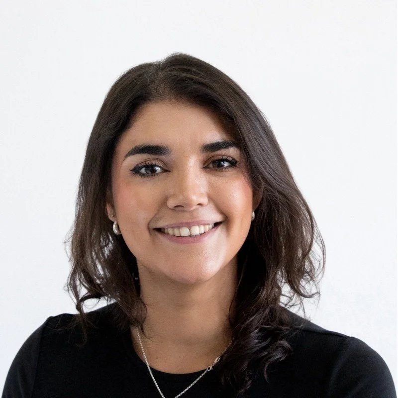 Ein Porträt einer jungen Frau mit dunklen, lockigen Haaren, die schwarze Kleidung trägt, vor hellem Hintergrund.