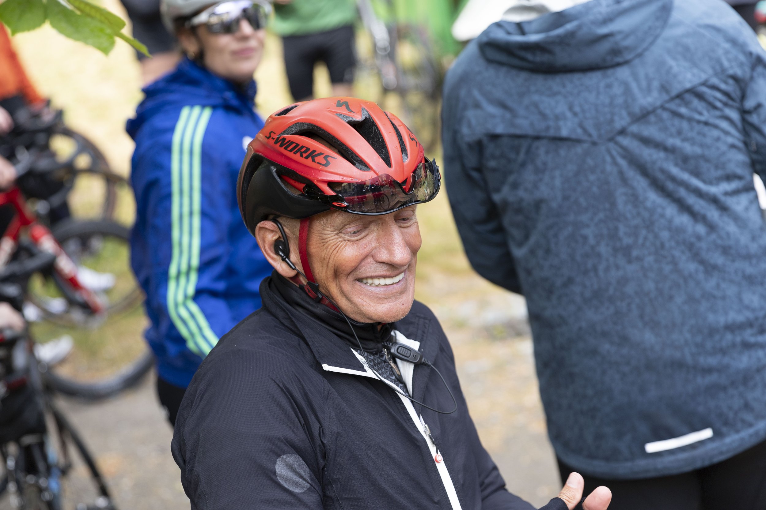 Ein lachender älterer Mann mit einem roten Fahrradhelm und einer schwarzen Jacke, offenbar bei einer Radfahrt im Freien, umgeben von anderen Radfahrern.