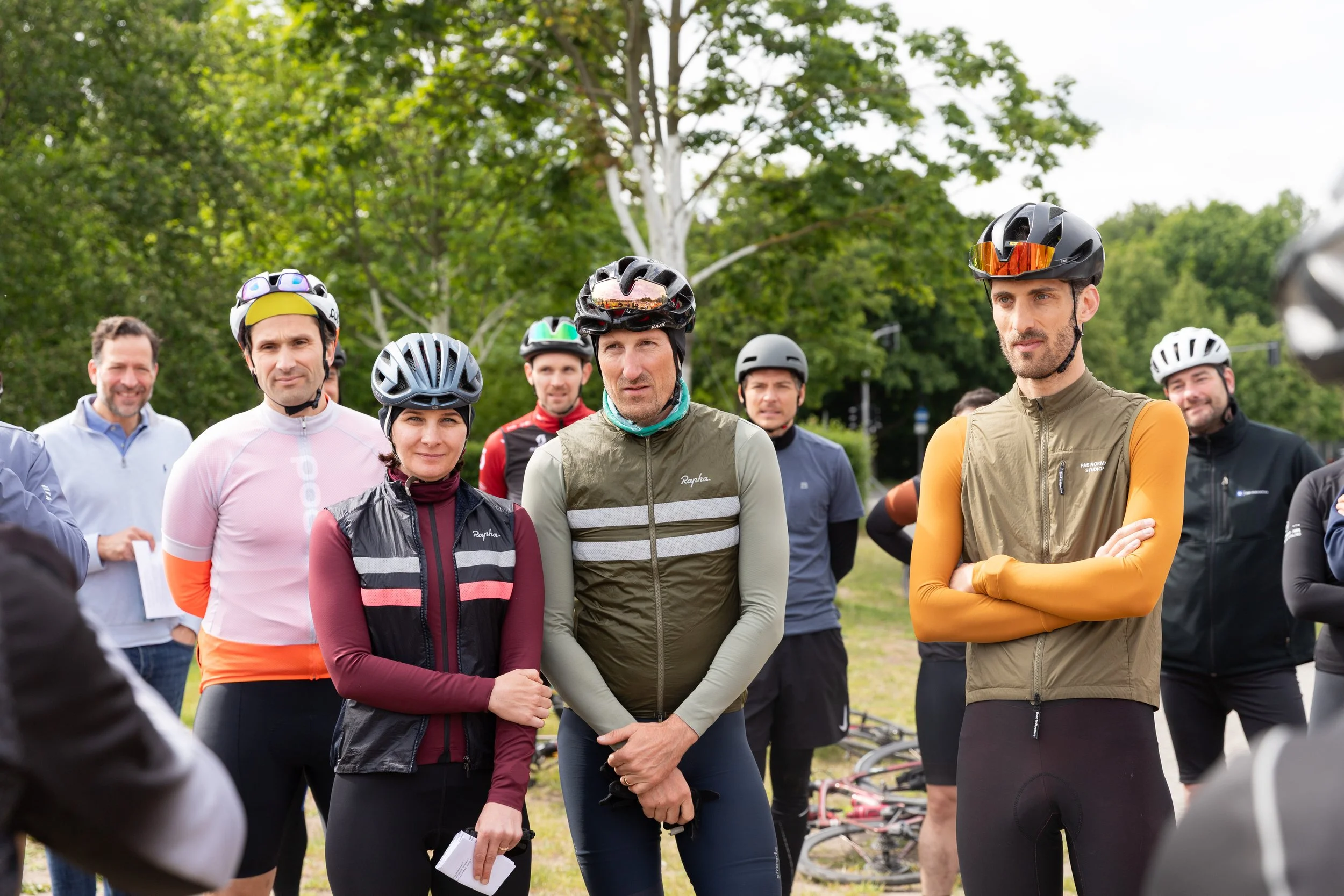 Eine Gruppe von Radfahrern in Helmen und Sportkleidung, die bei einem Treffen im Freien zuhört.