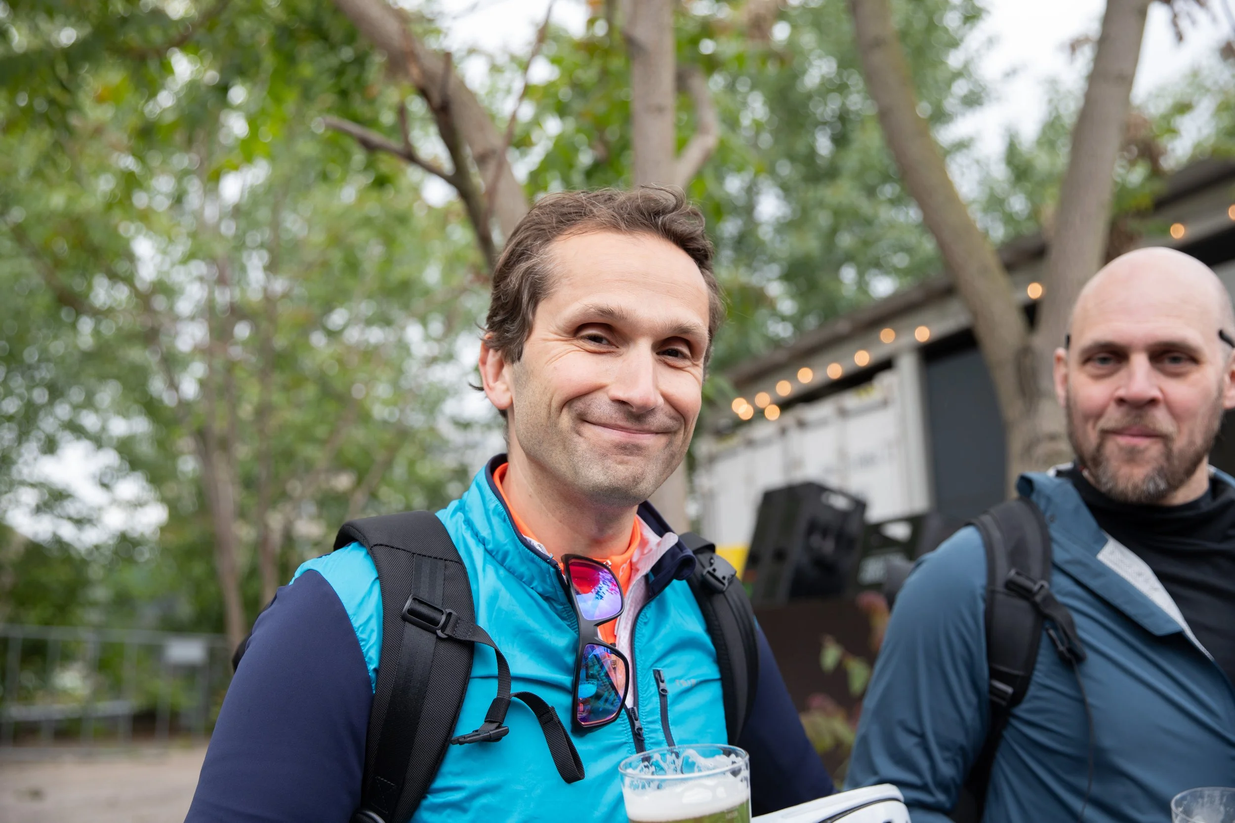 Zwei Männer mit Rucksack beim Outdoor-Event, einer hält ein Glas Bier, die Umgebung ist grün mit Bäumen, im Hintergrund Lichterketten und ein Gebäude.