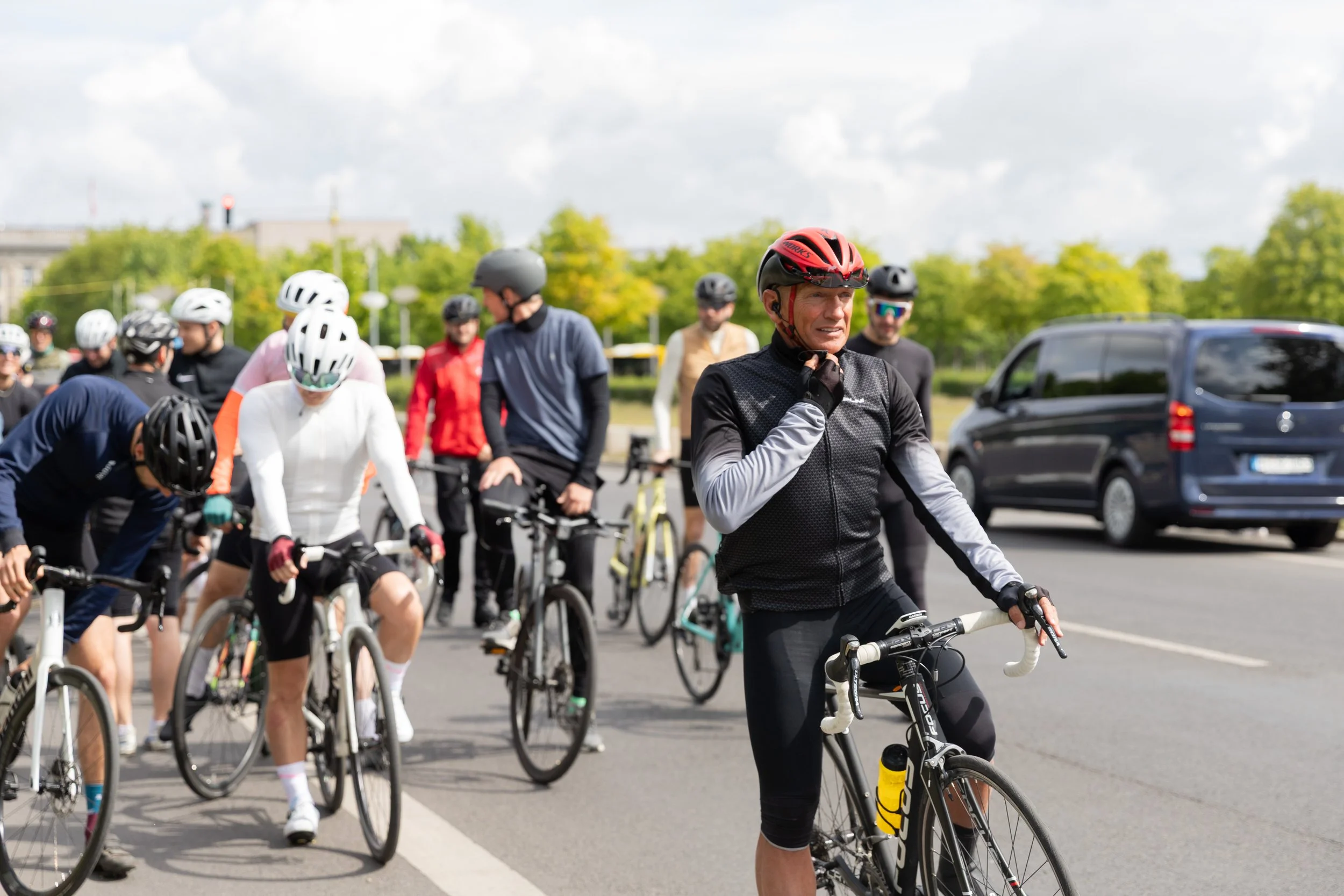Gruppe von Radfahrern am Straßenrand, einige sind mit Helmen, in sportlicher Kleidung, während ein Mann im Vordergrund, auch mit Helm, auf einem Rennrad steht und nachdenkt.