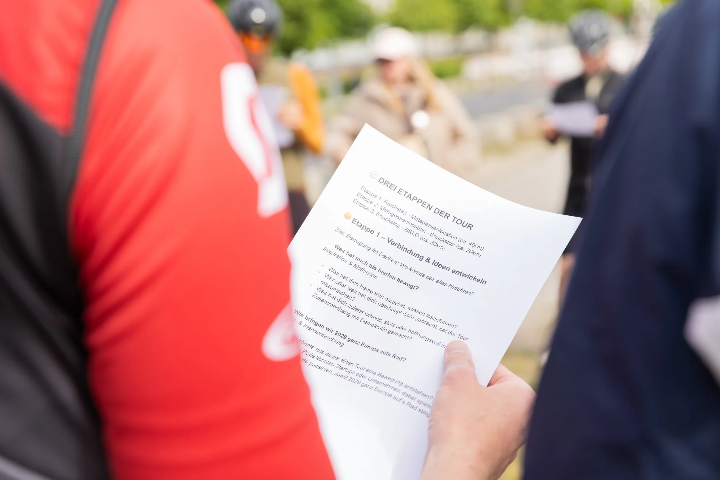 Person hält einen Flyer mit Tour-Informationen, während im Hintergrund mehrere Menschen stehen, einige mit Helmen, im Freien.