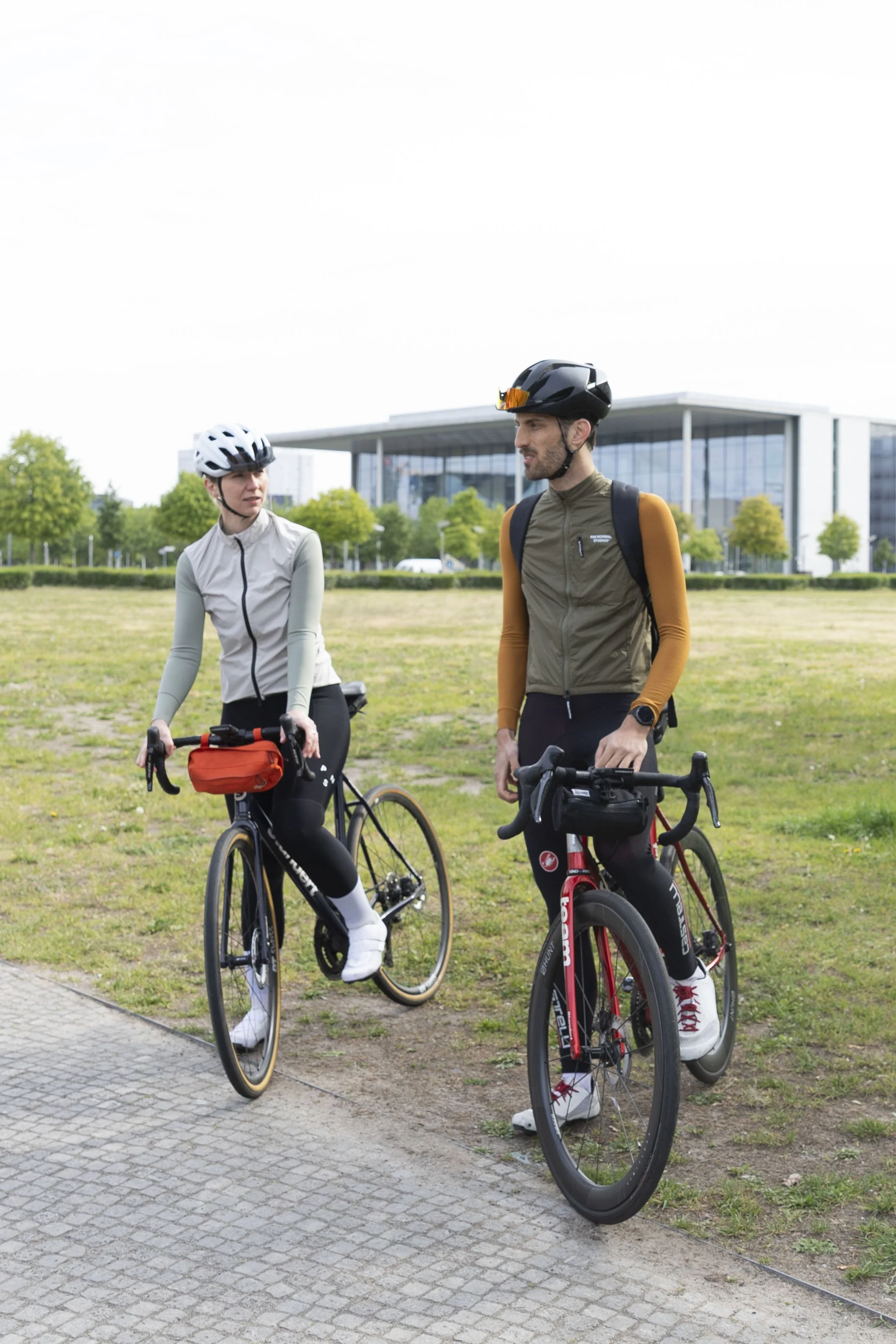 Zwei Radfahrer sprechen im Park, beide tragen Helme und rüsten sich für eine Fahrradtour, im Hintergrund moderne Gebäude.