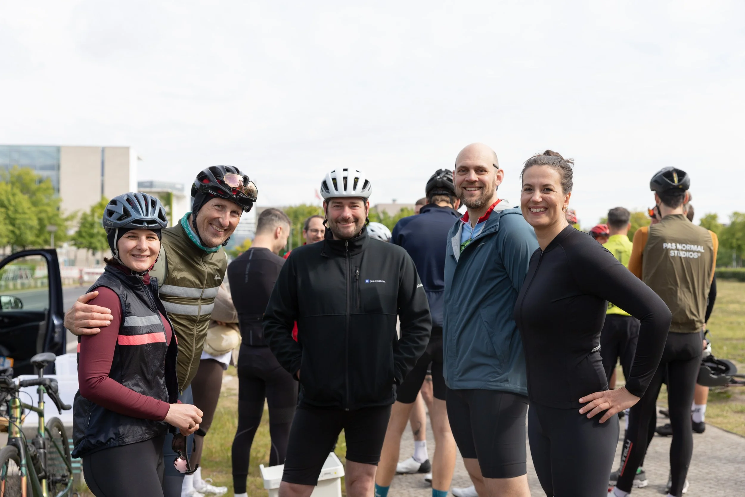 Fünf Menschen in Fahrradhelmen und Sportkleidung beim Gruppenfoto im Freien, während andere Radfahrer im Hintergrund bereitstehen.