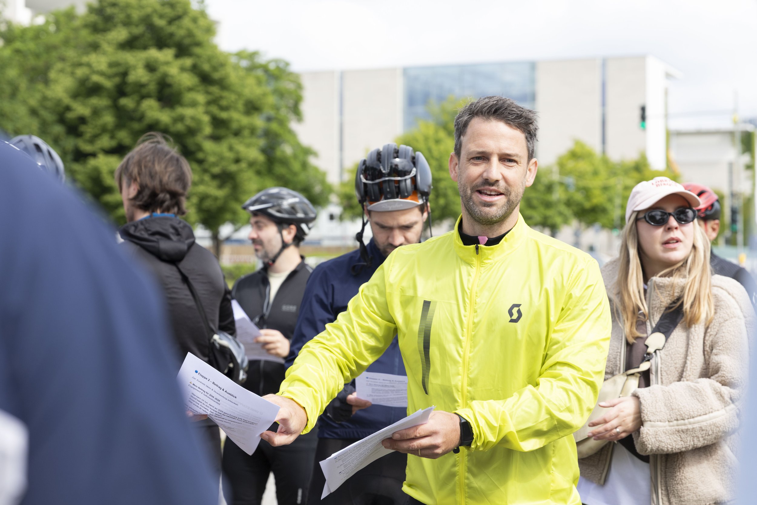 Mann in gelber Jacke verteilt Flyer an Fahrradfahrer bei einer Gruppenveranstaltung im Freien.