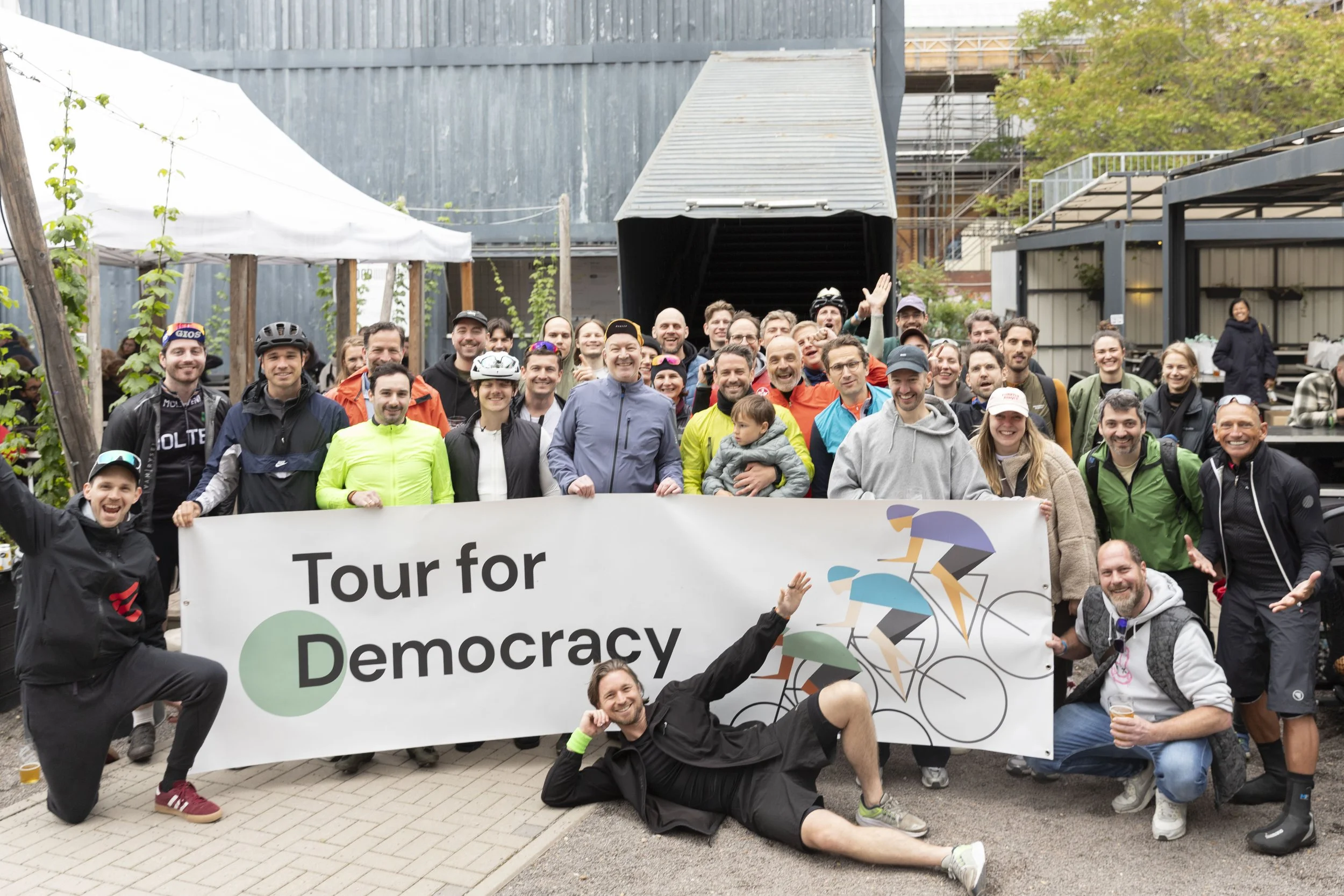 Gruppe von Menschen bei einer Fahrradtour, die ein Banner mit der Aufschrift 'Tour for Democracy' halten.