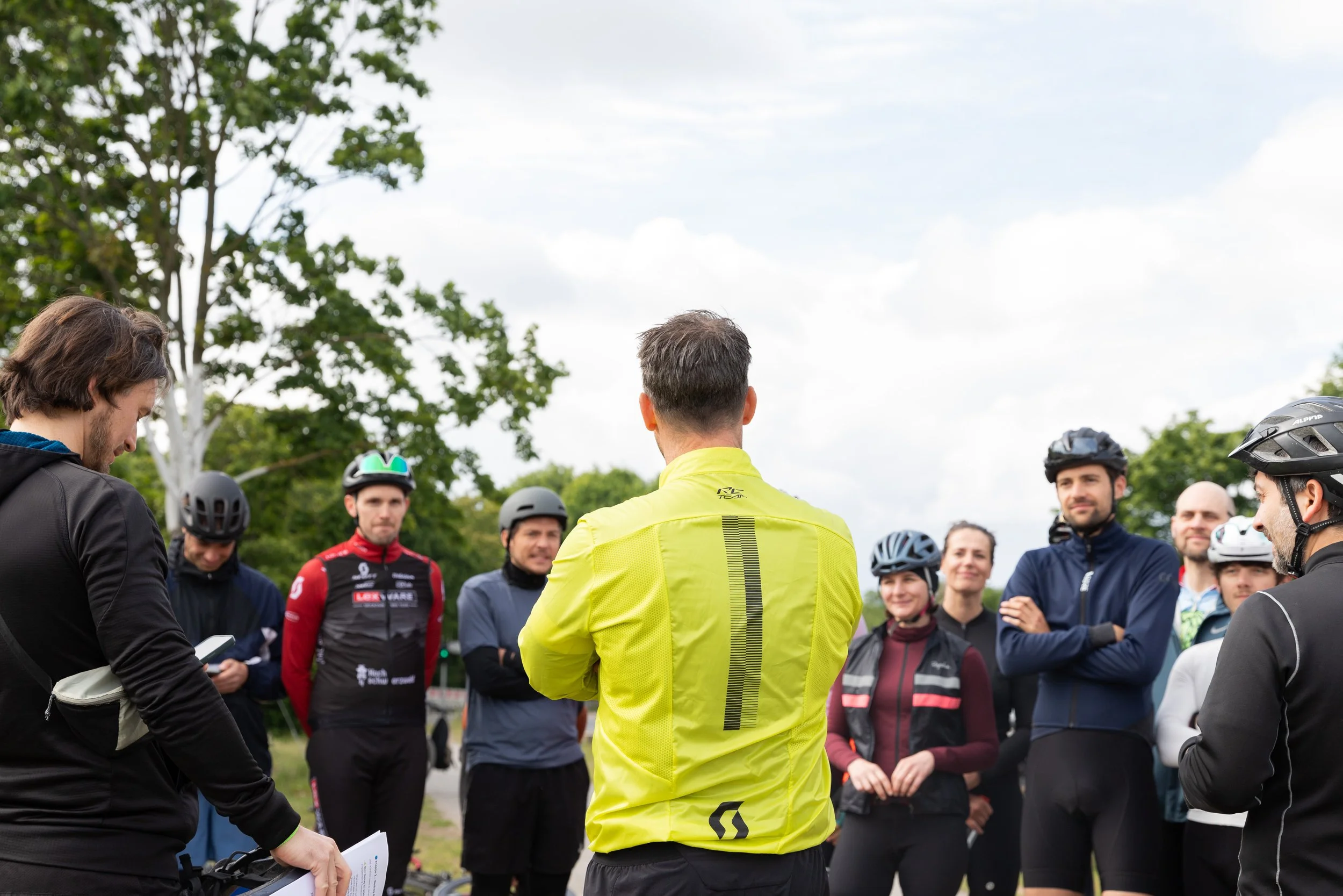 Gruppe von Menschen, die in einem Park stehen, einige tragen Fahrradhelme, während ein Mann in einer leuchtend gelben Jacke spricht.