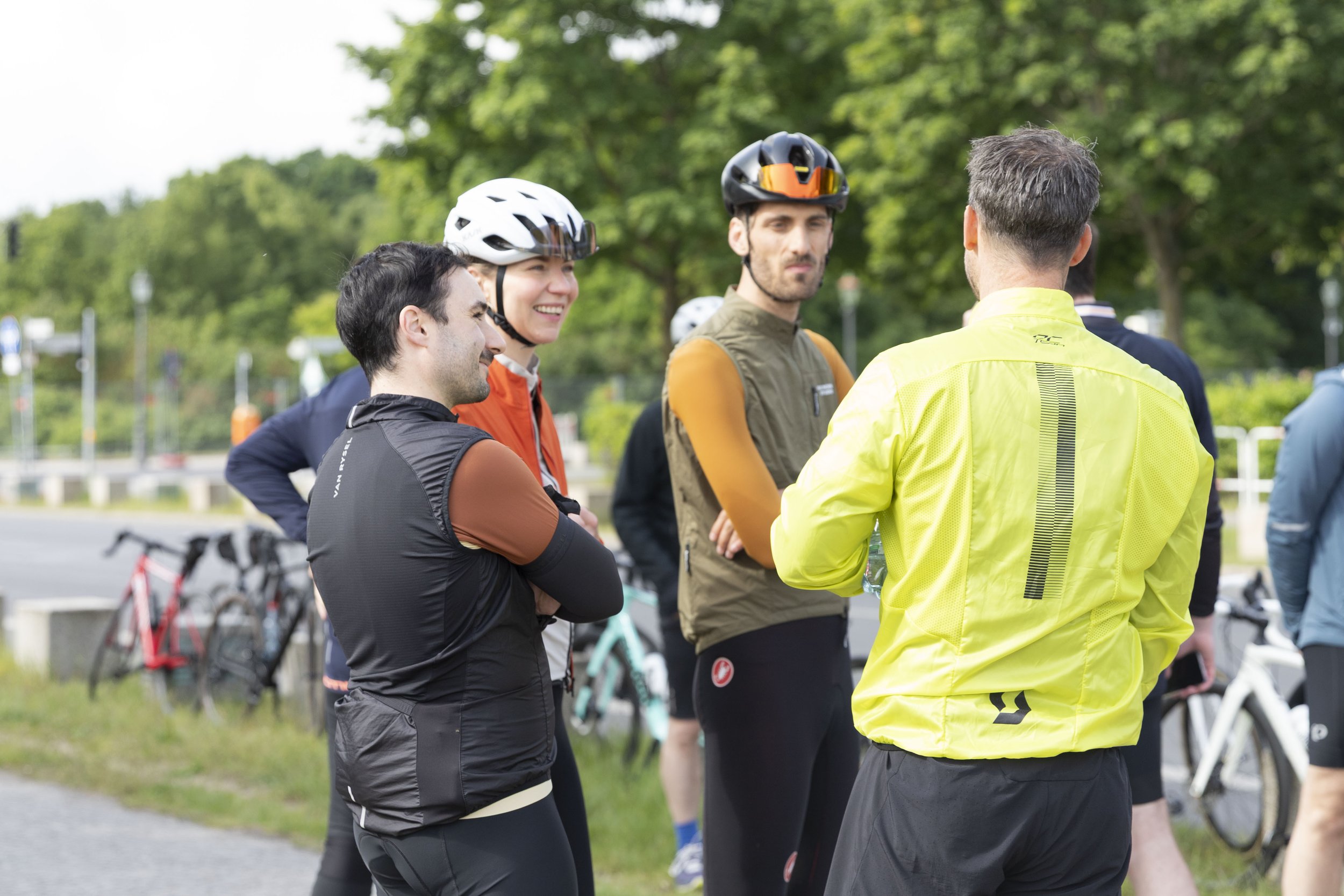 Eine Gruppe von Radfahrern, die im Gespräch stehen, einige tragen Helme und Fahrradkleidung, im Hintergrund sind Fahrräder und Bäume.