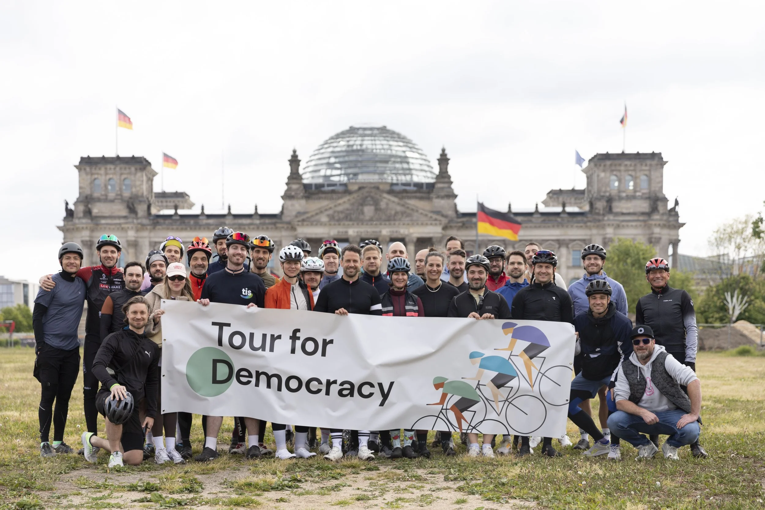 Gruppenfoto von Radfahrern vor dem Bundestagsgebäude mit einem Banner, auf dem steht 'Tour for Democracy' mit Illustrationen von Radfahrern.