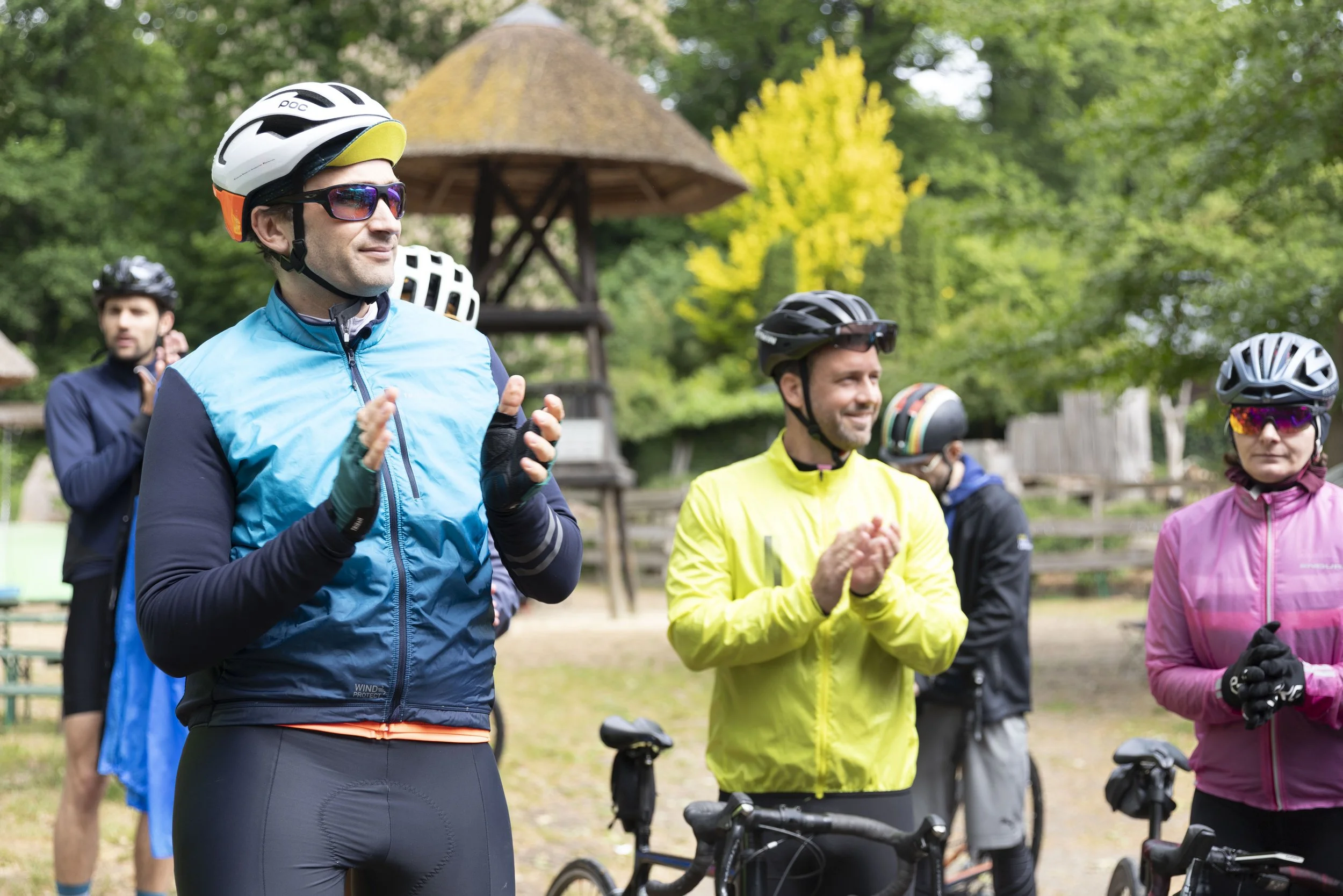 Gruppe von Radfahrern in Fahrradausrüstung, die draußen im Grünen stehen, applaudierend und lächelnd.
