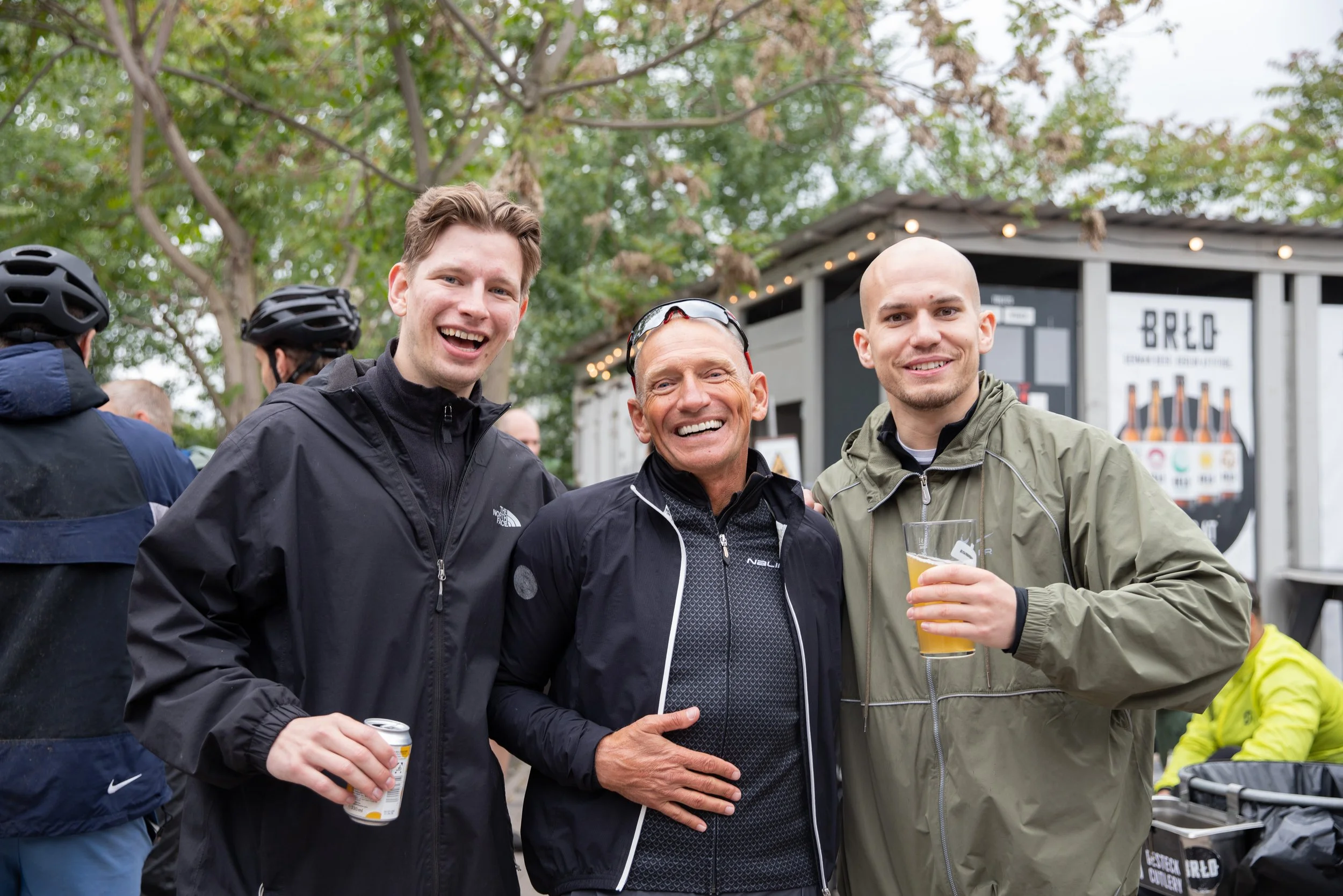 Drei Männer auf einem Fest, sie lachen und halten Getränke, im Hintergrund sind andere Menschen und eine kleine Bude.