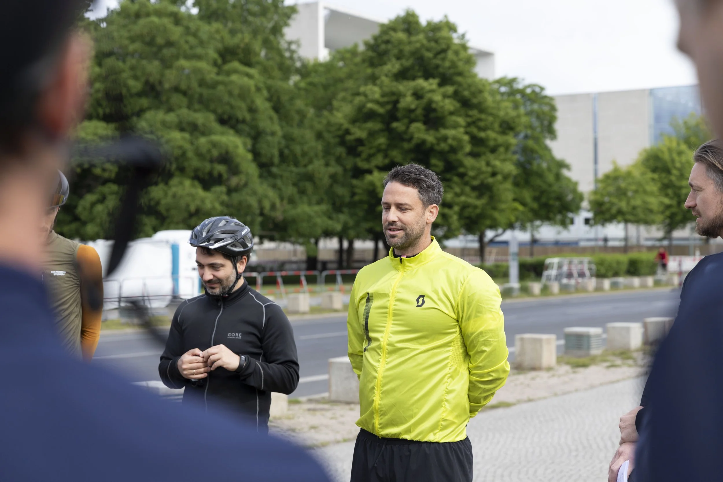 Eine Gruppe von Menschen, einige in Sportkleidung, trifft sich outdoors auf einem Weg neben einer Straße, im Hintergrund grüne Bäume und moderne Gebäude.