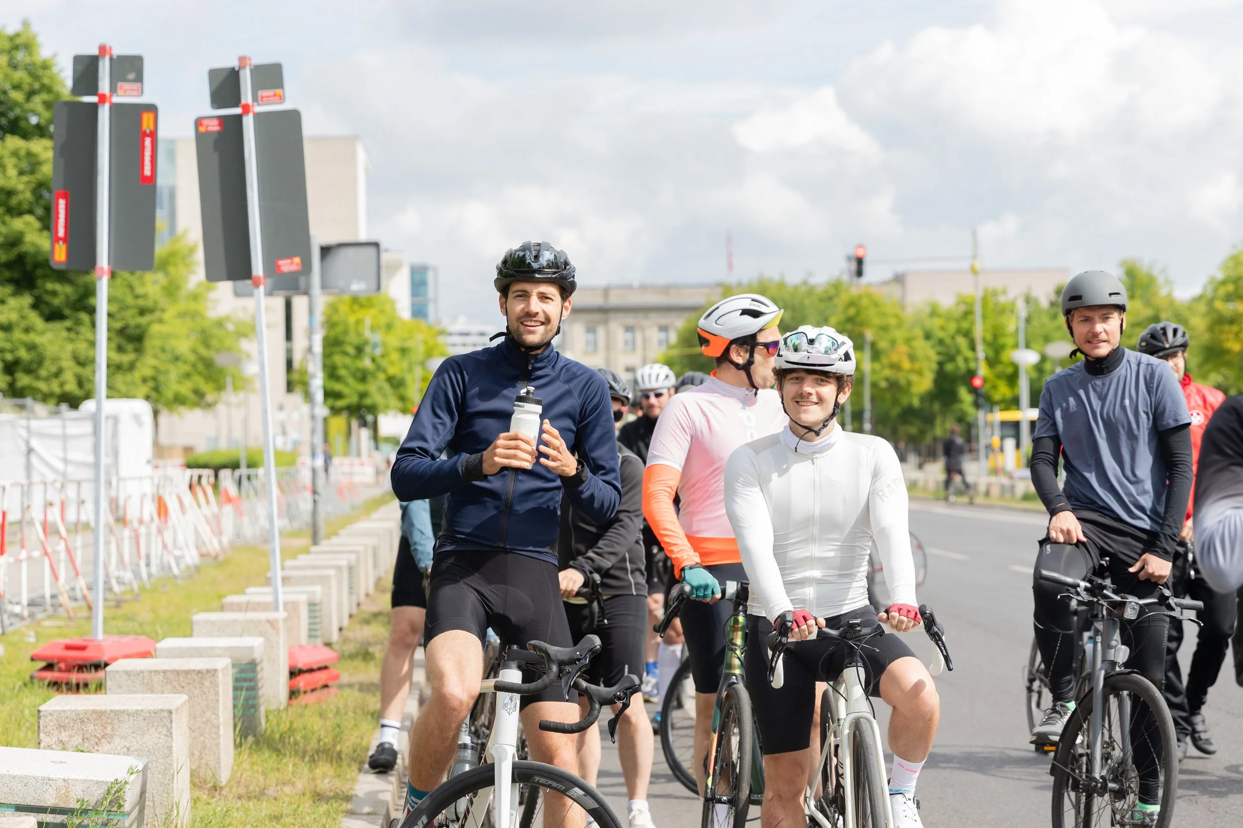 Gruppe von Radfahrern, die auf einer städtischen Straße stehen, alle mit Helmen, einige lächelnd und sich unterhaltend, während sie sich auf ihre Fahrräder vorbereiten.