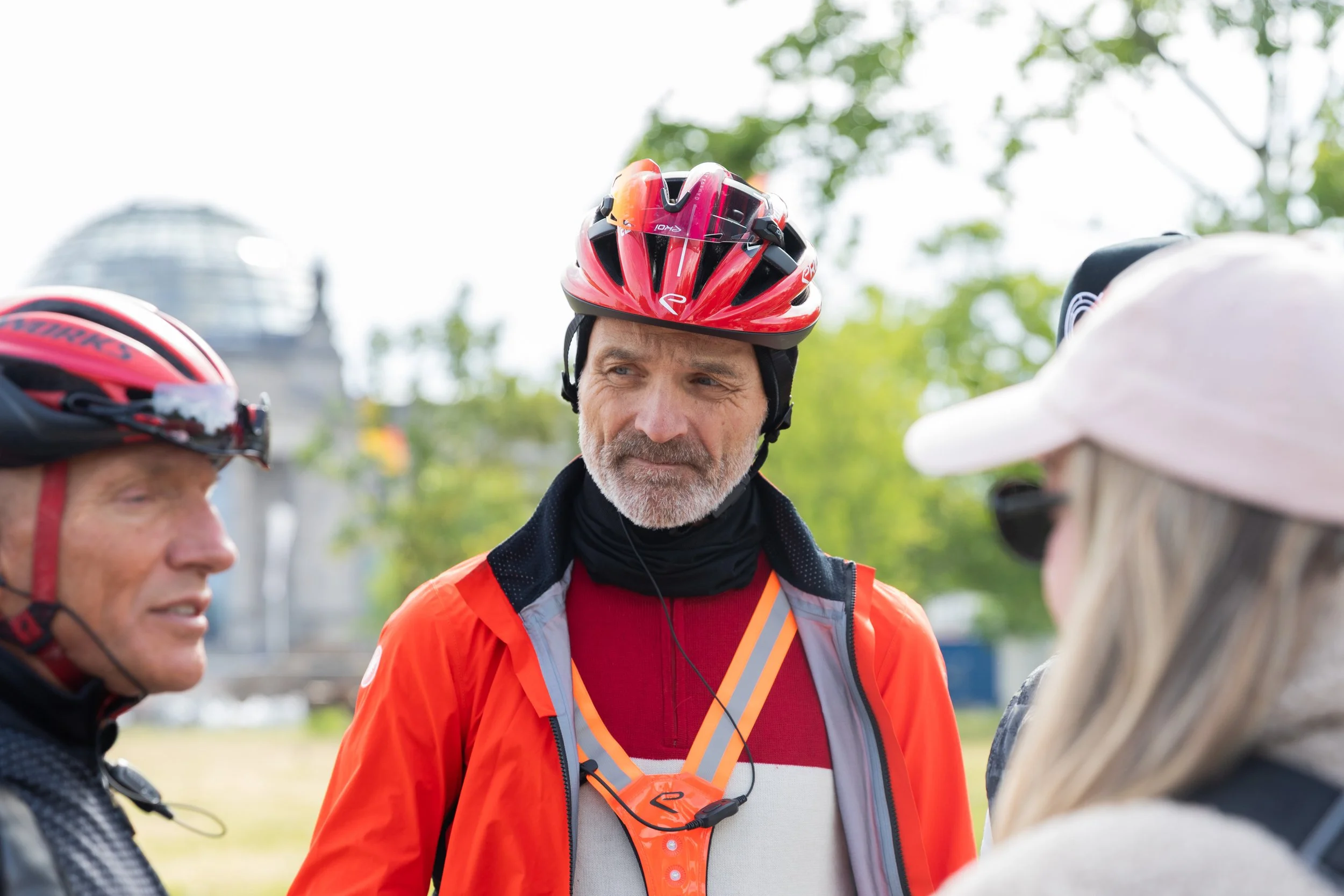 Drei Fahrradfahrer im Gespräch, einer trägt einen roten Helm, der andere einen schwarzen Helm, im Hintergrund Bäume und ein Gebäude.