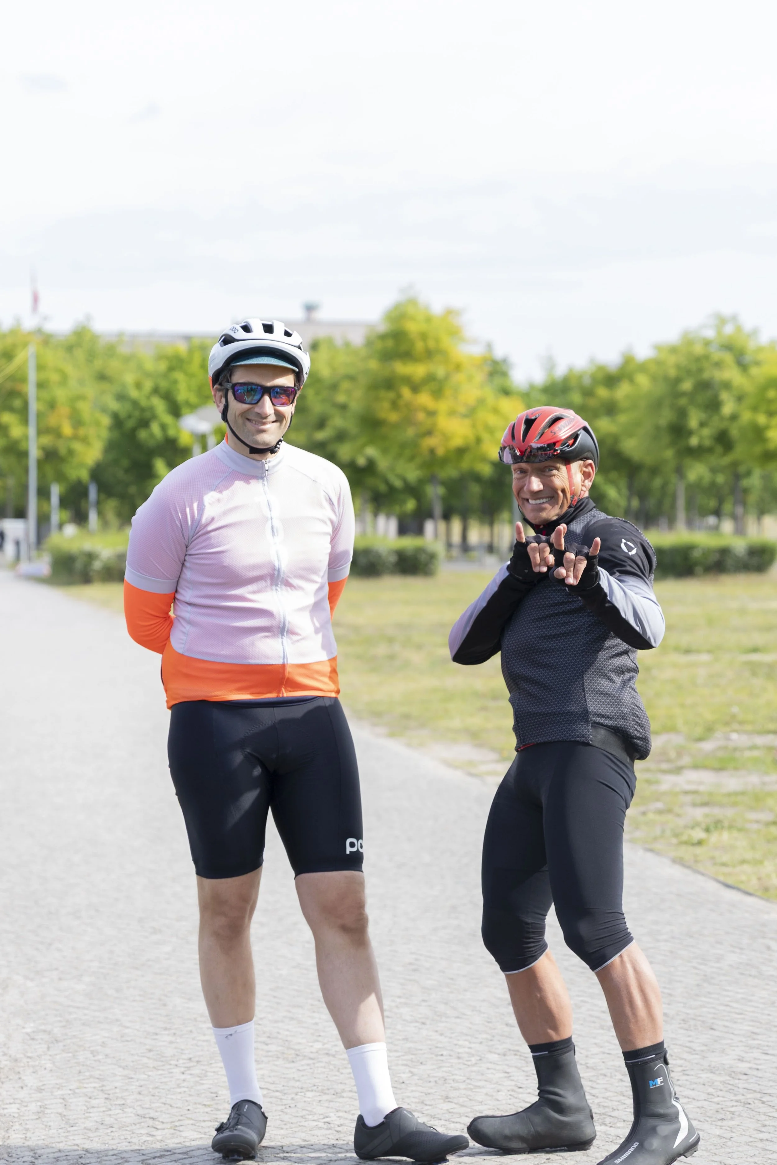 Zwei männliche Radfahrer in Sportkleidung und Helmen stehen auf einem Weg im Park, einer lächelt in die Kamera, der andere macht die Handzeichen für 'I love you'.