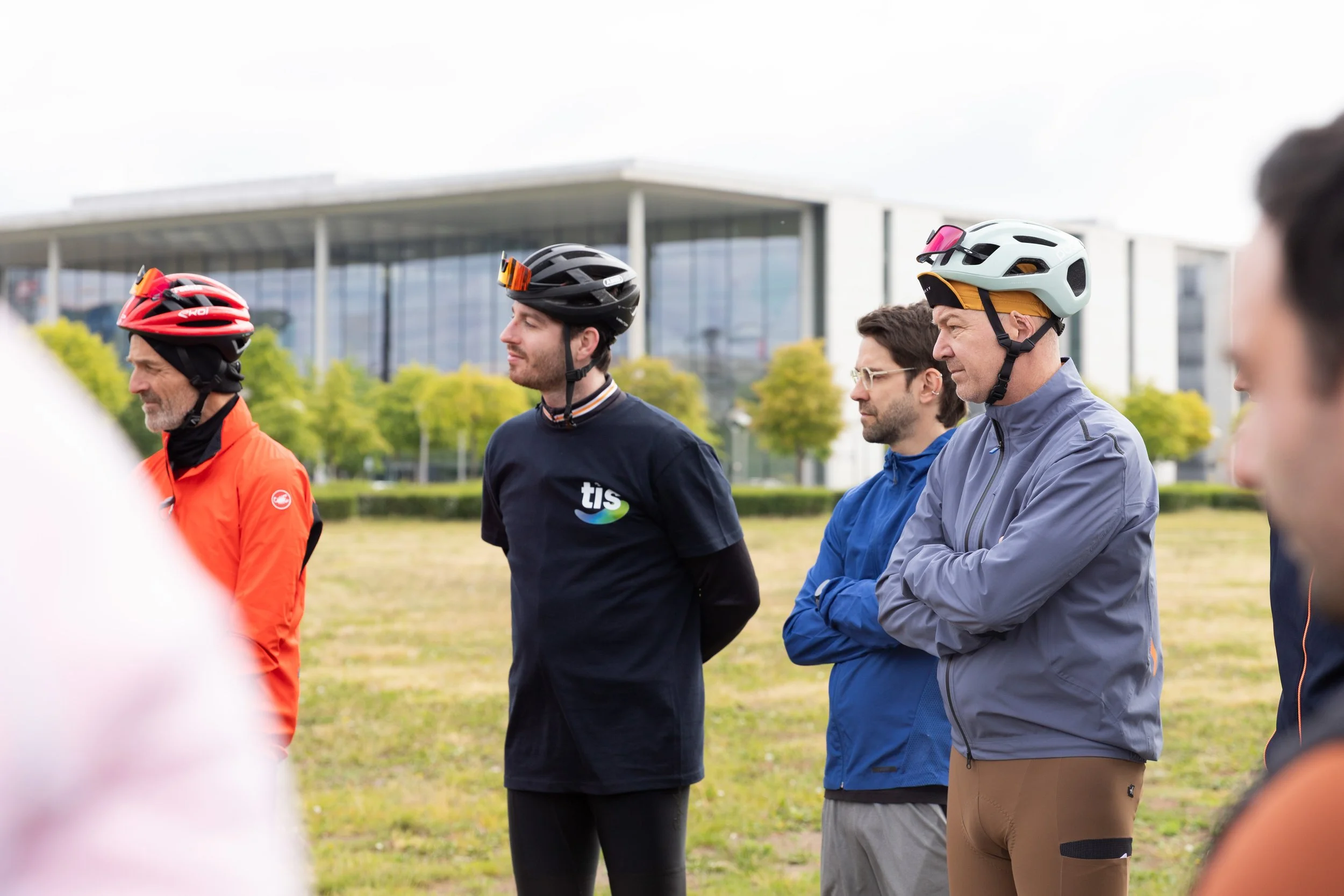 Fünf Menschen mit Fahrradhelmen stehen im Freien in einer Reihe, während sie einem Sprecher zuhören, mit moderner Gebäudearchitektur und Bäumen im Hintergrund.