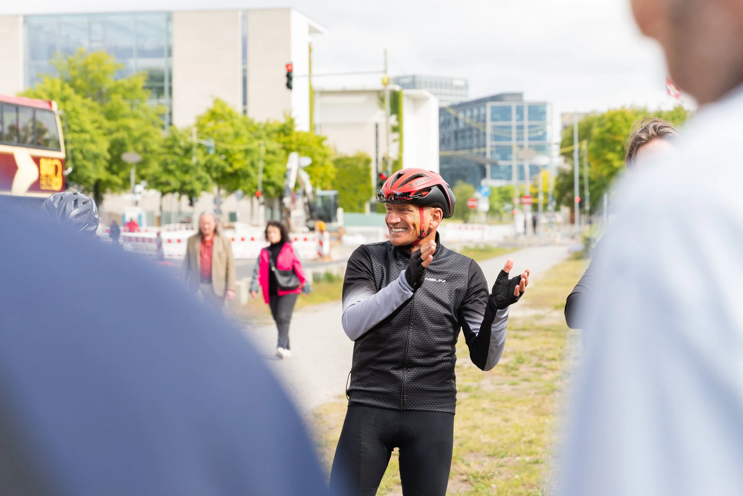 Ein männlicher Radfahrer trägt einen Helm und eine Fahrradjacke, lächelt und klatscht, während er mit anderen Menschen im Freien spricht, mit modernen Gebäuden und grünen Bäumen im Hintergrund.