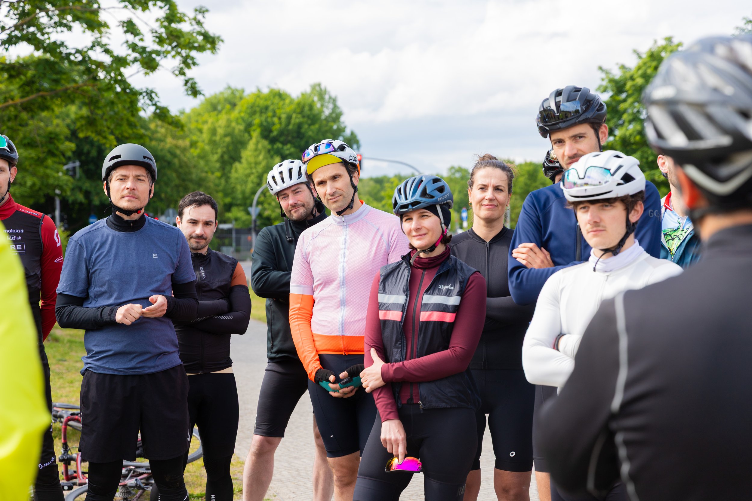 Gruppe von Radfahrern im Gespräch, alle tragen Fahrradhelme und Sportkleidung, im Hintergrund Bäume und ein Himmel mit Wolken.