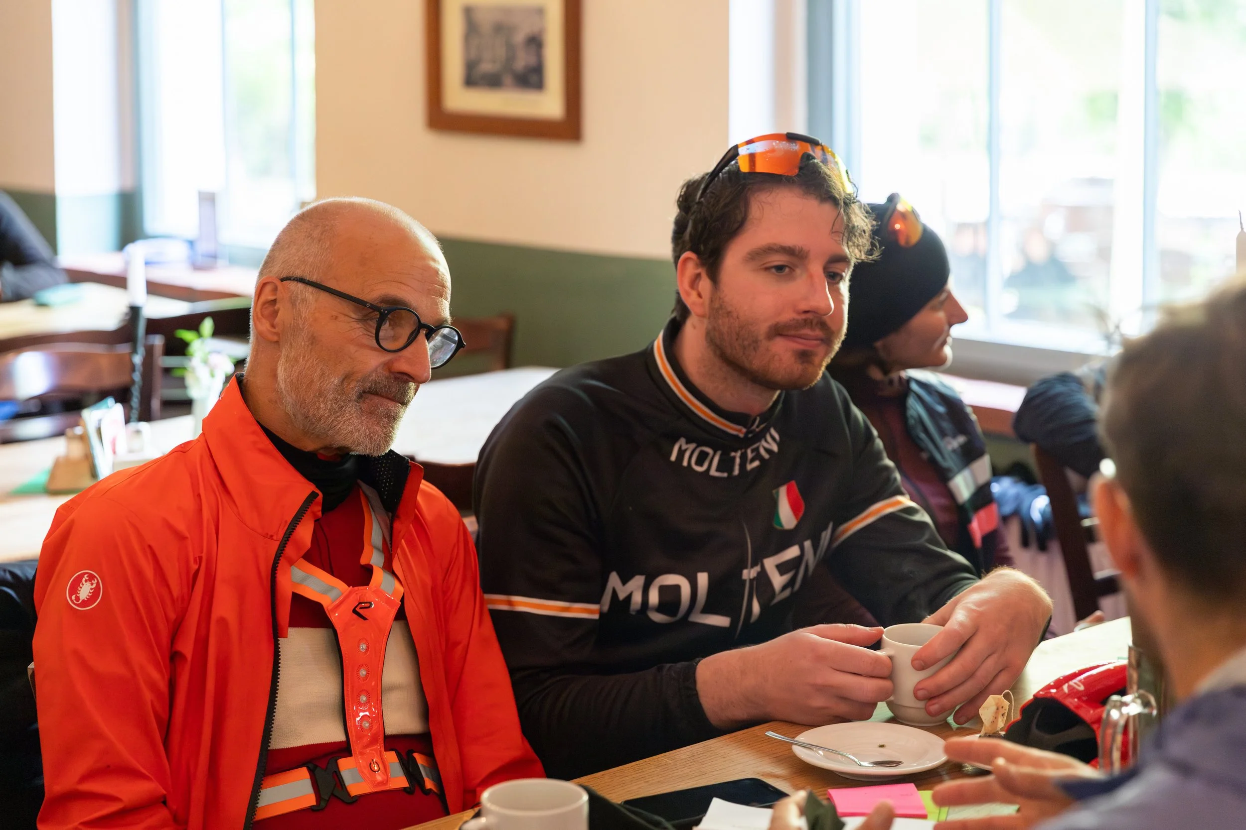 Drei Personen sitzen in einem Café, trinken Kaffee. Der Mann in der Mitte trägt eine schwarze Jacke mit Markenlogo, der Mann links trägt eine orange Fahrradjacke.