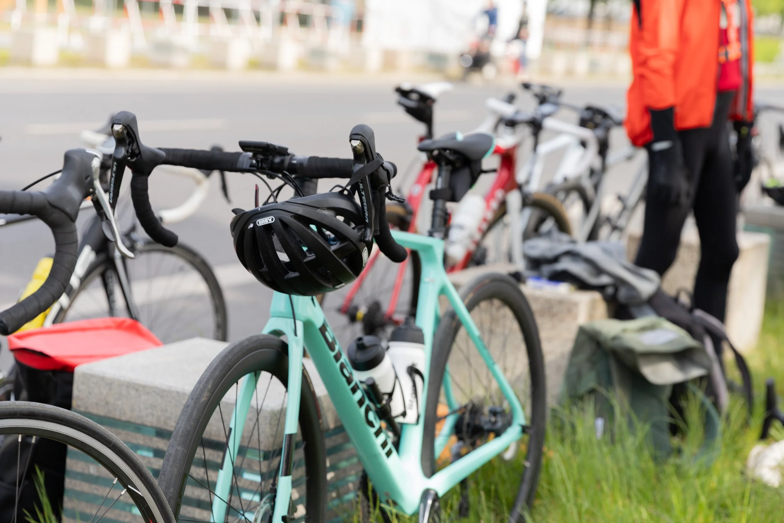 Mehrere Fahrräder, darunter ein kleines, grün-blaues Fahrrad mit Schutzhelm an einem Sitzgitter, stehen am Straßenrand. Im Hintergrund sind Personen, die sich in einem Fahrradparkbereich aufhalten.