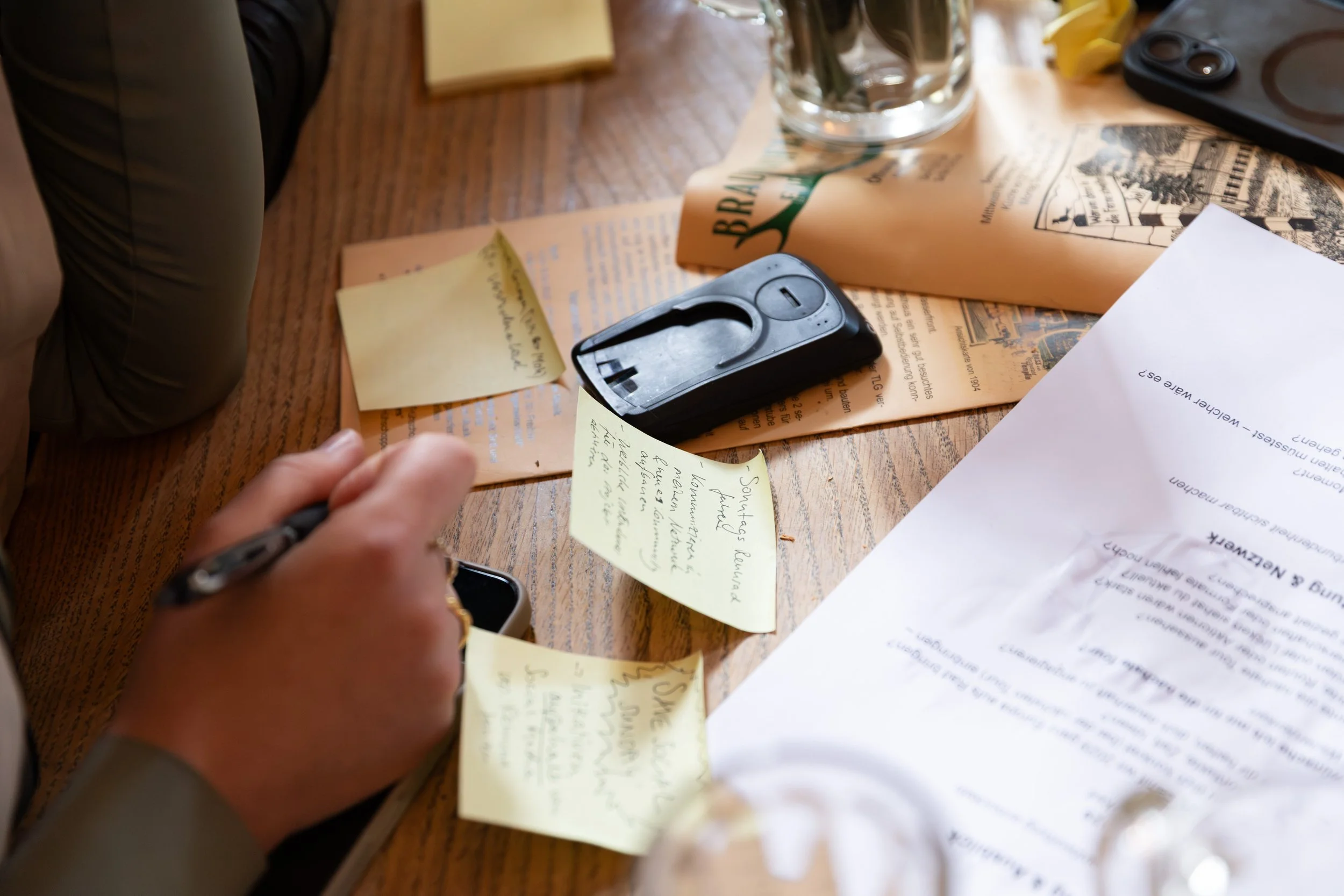 Ein Tisch mit mehreren Stückchen Papier, Notizzettel, einem Handy, einem Taschenrechner, einer Glasvase mit Blumen, Bananen und weiteren Gegenständen.