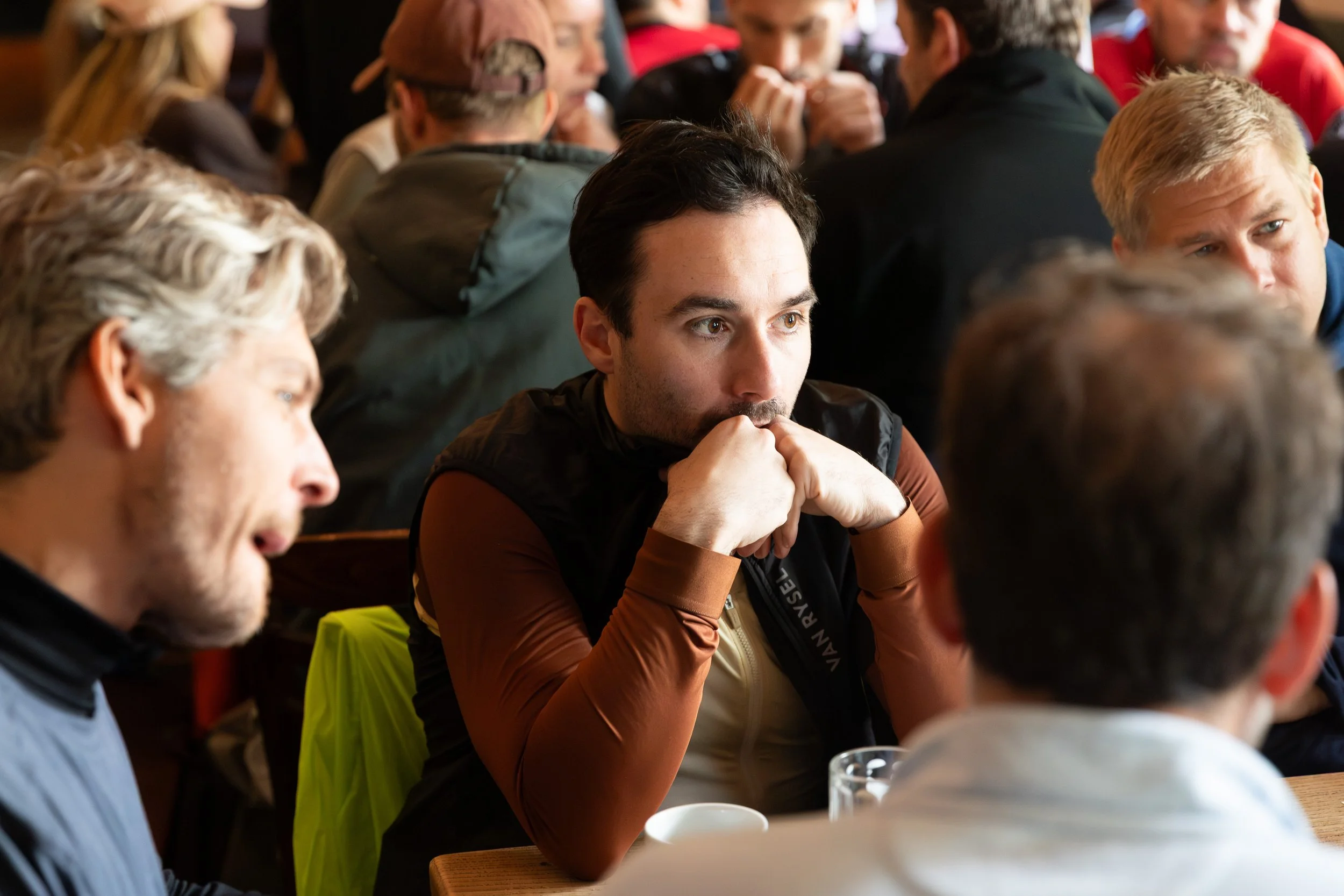 Mehrere Menschen sitzen in einem Raum, einige betrachten nachdenklich oder konzentriert. Im Vordergrund befindet sich eine Frau mit dunklen Haaren, die den Blick nach unten richtet, während ein Mann mit braunen Haaren und einer schwarzen Weste auf ih