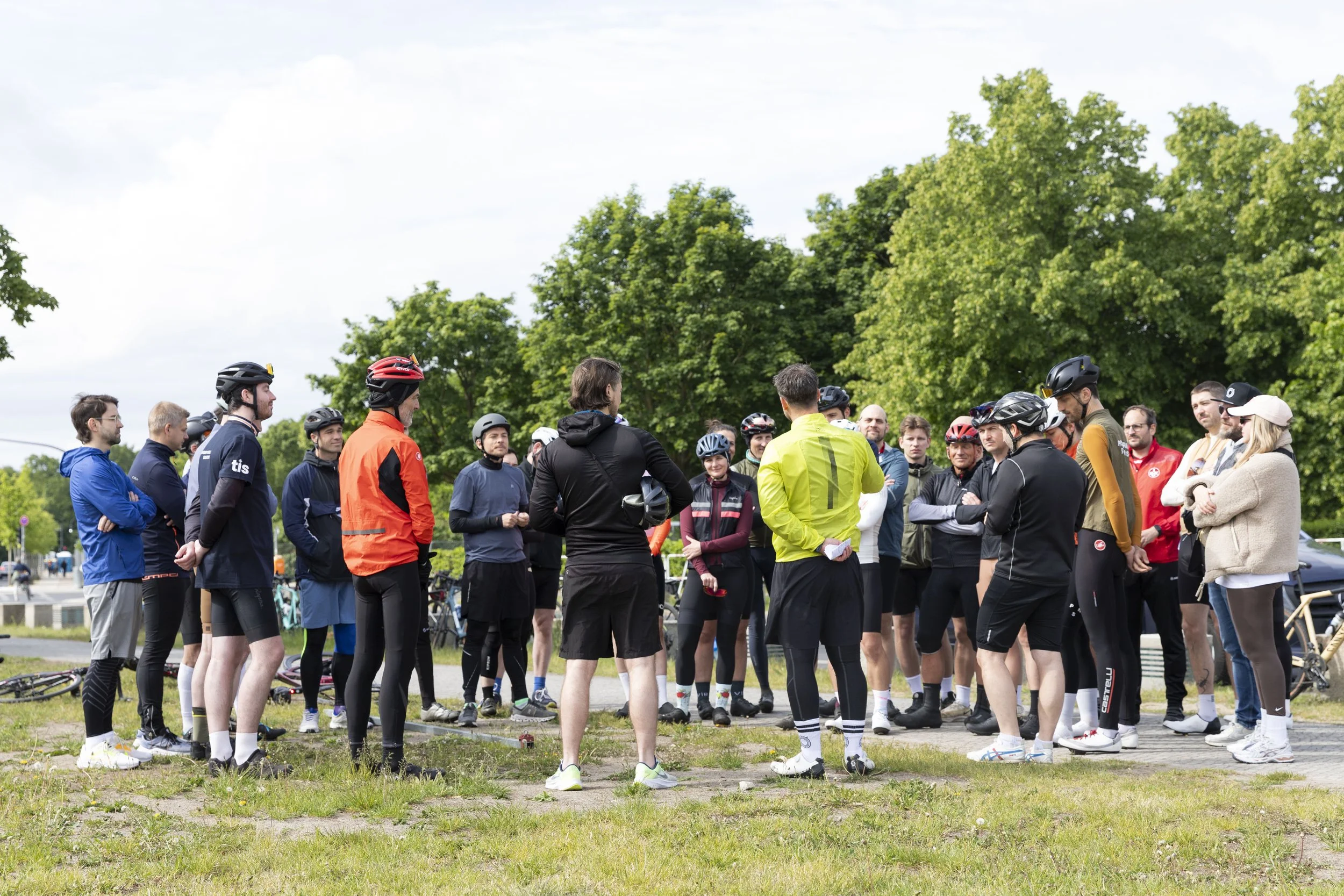 Gruppe von Radfahrern und Läufern, die im Freien auf einer Wiese versammelt sind, während eine Person in Gelb vorredet.