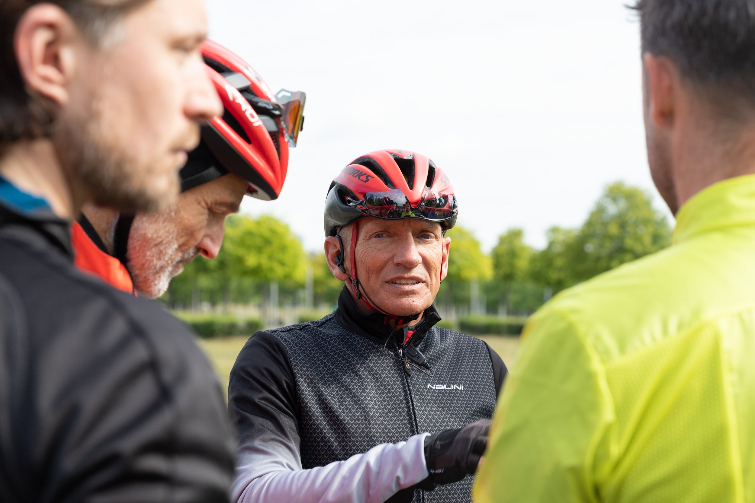 Vier Menschen mit Fahrradhelmen und Kleidung, die wahrscheinlich Fahrradfahrer sind, stehen in einer Diskussion im Freien, mit Bäumen im Hintergrund.