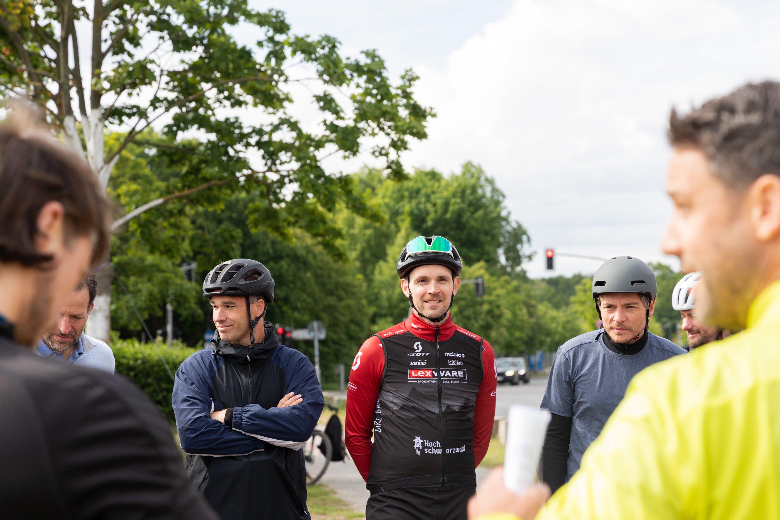 Mann in rotem Fahrradtrikot spricht mit Gruppe von Männern, einige tragen Helme, im Freien bei grünem Baum.