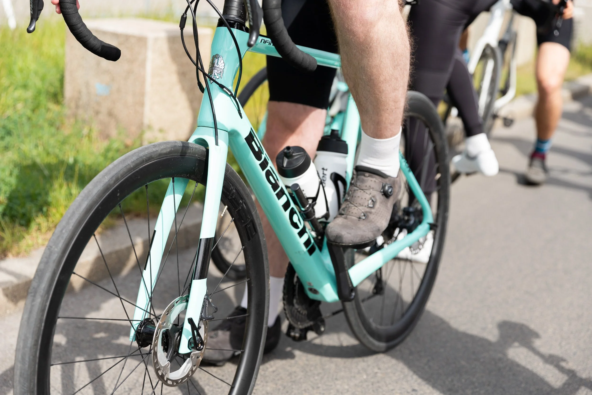 Fokus auf das Vorderteil eines hellblauen Rennrads mit dem Namen 'Bianchi', das von einem Radfahrer mit grauen Socken und Sportschuhen benutzt wird. Im Hintergrund sind weitere Radfahrer zu sehen.